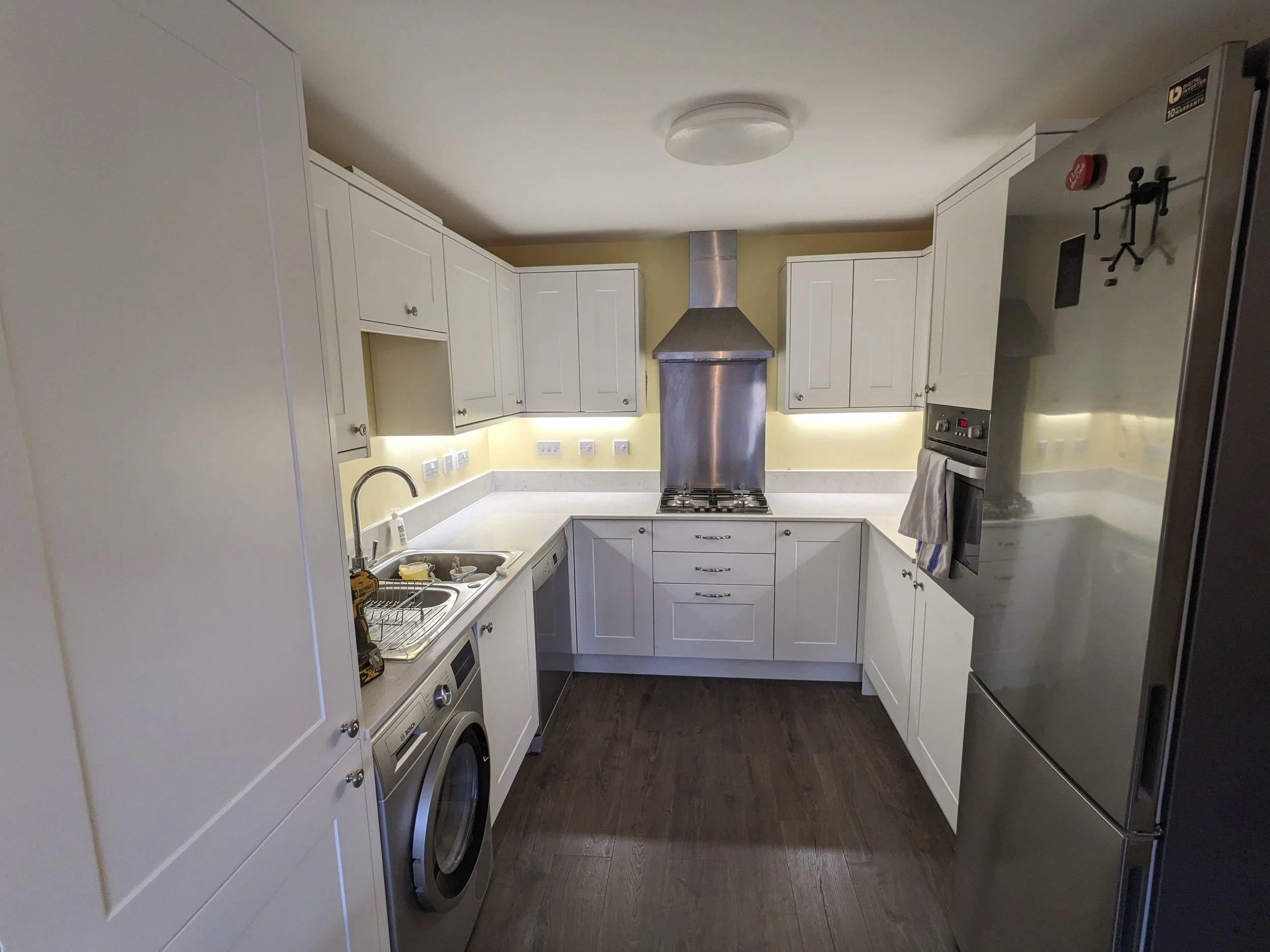A small L-shaped kitchen with white cabinets, a stainless steel range hood, and a dark wood floor. Includes a sink, washing machine, and built-in oven.