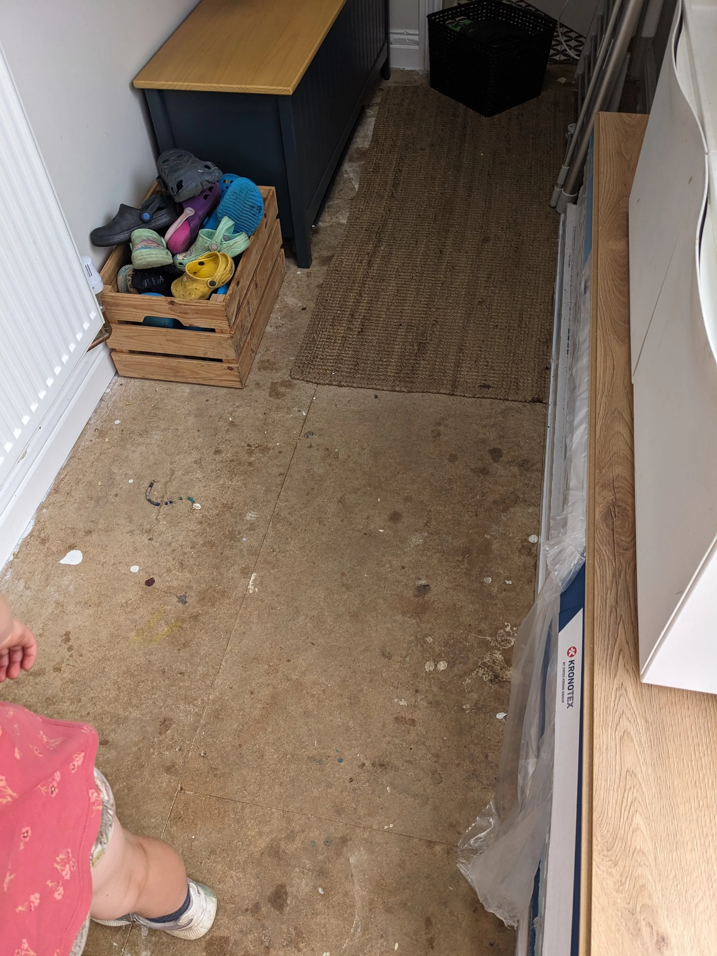 A small entryway or mudroom with a beige floor, brown doormat, and various storage items, including a wooden crate filled with children's shoes, a dark blue cabinet, a black basket, and a small table with storage drawers. Part of a child's leg and sh