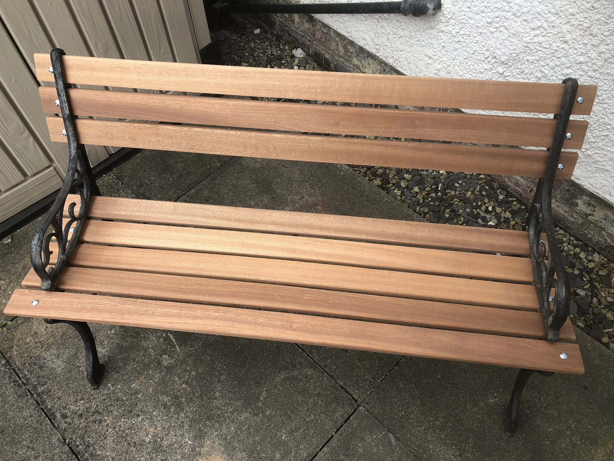A wooden outdoor bench with a black metal frame is placed on a stone patio next to a white wall and a gray shed.