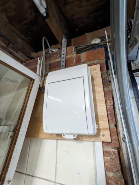 A white electrical box mounted on a wooden board on a brick wall in a garage or workshop.