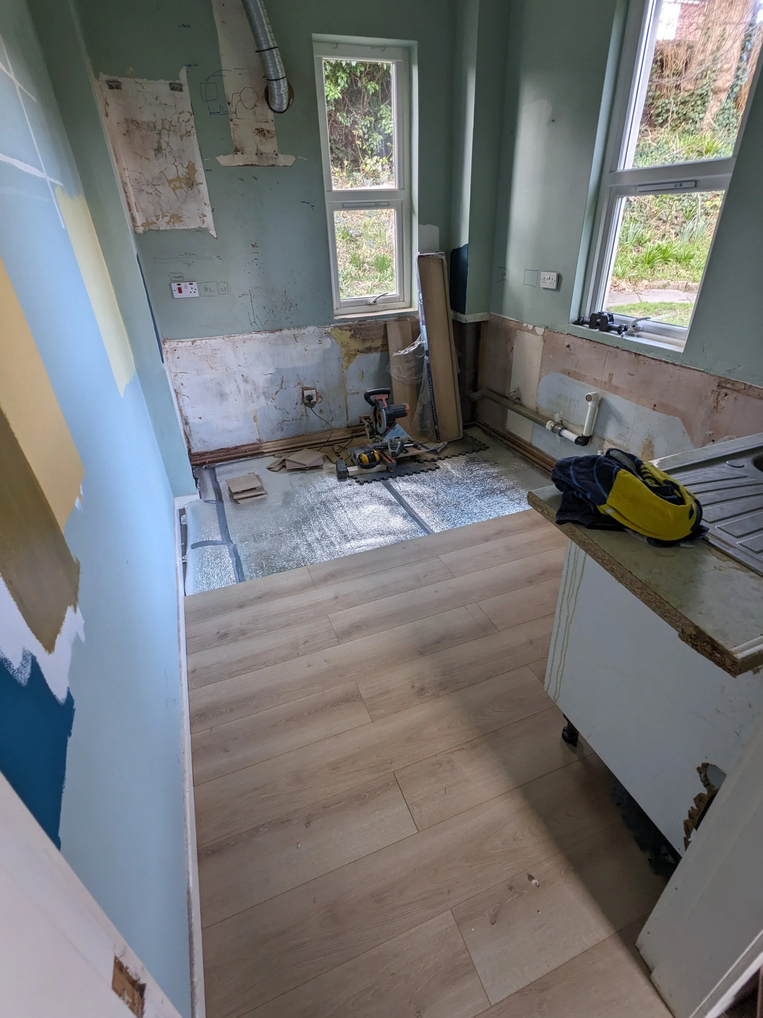 A partially renovated kitchen with new light wood flooring, green walls, and two windows showing greenery outside. The lower part of the walls is unfinished, with exposed pipes and drywall, and construction tools are on the floor.
