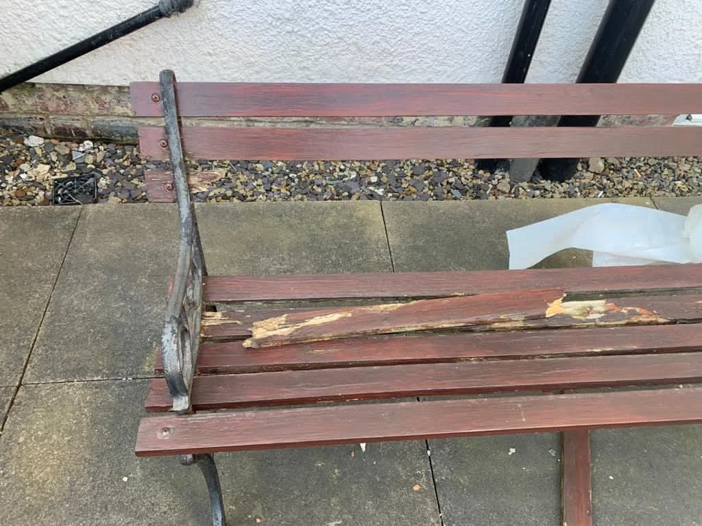 A wooden park bench with a metal frame, showing signs of wear and peeling paint, on a concrete sidewalk next to a building wall and gravel area.