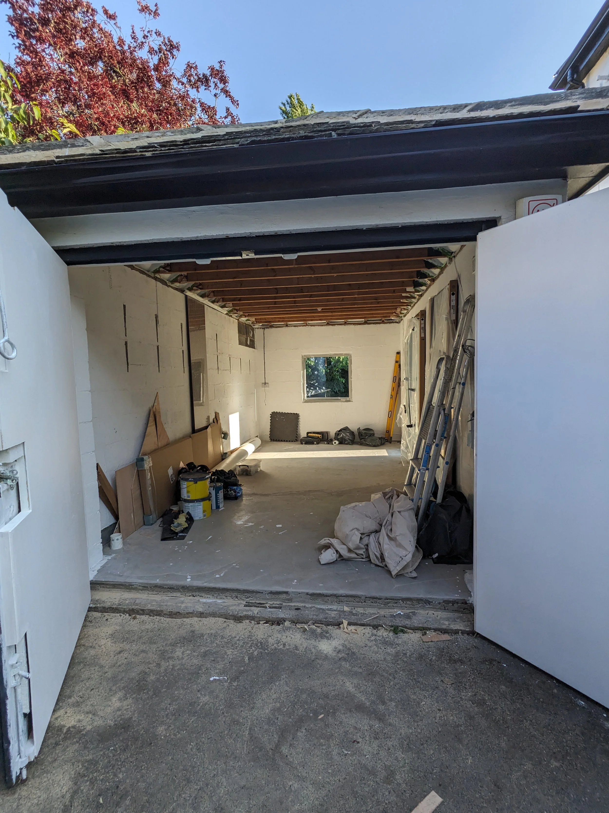 View inside a garage under renovation, showing construction tools, ladders, paint cans, and construction materials with an unfinished ceiling and a small window at the end.