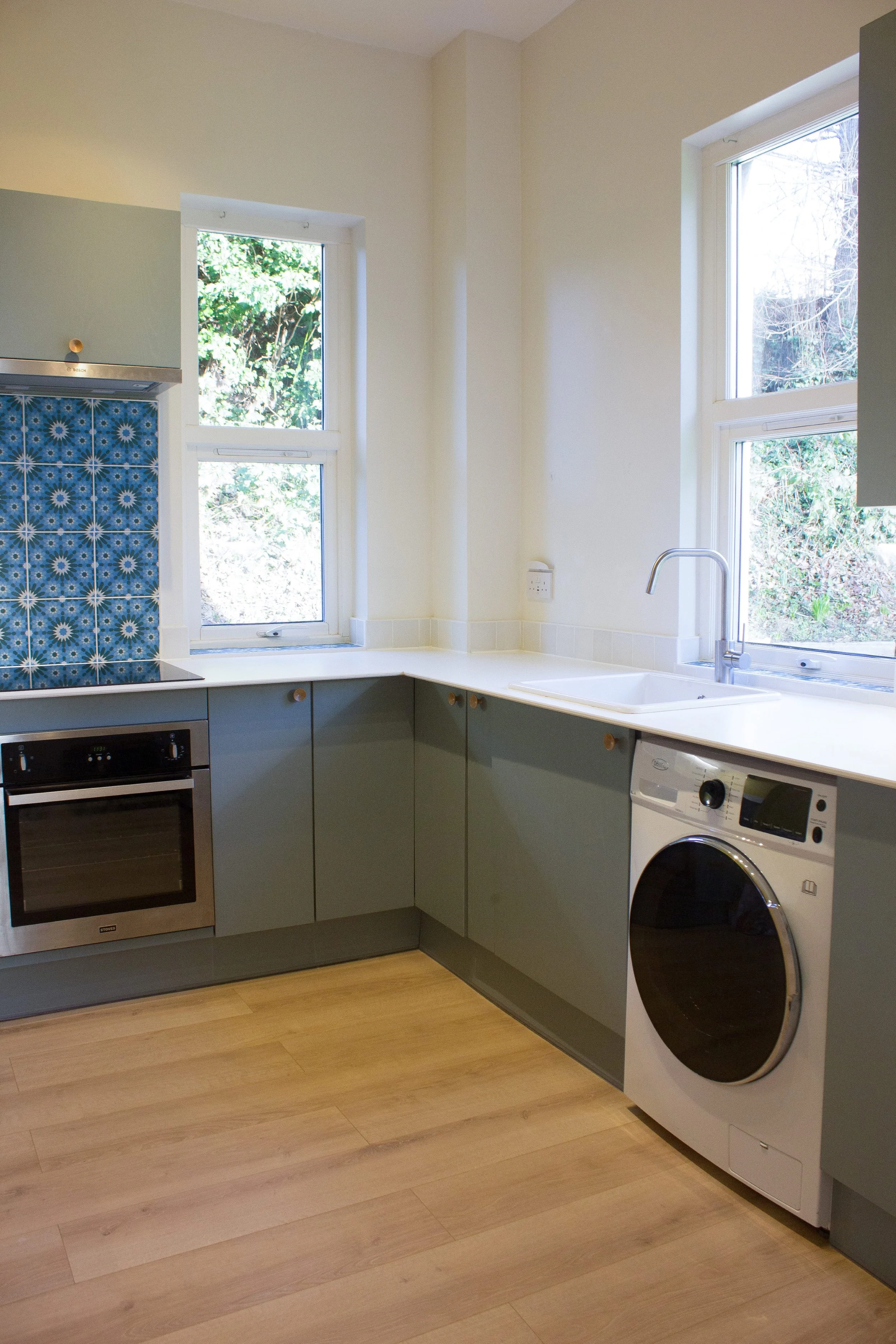 A kitchen with light green cabinets, a washing machine next to the sink, a built-in oven, two windows with natural light, a blue patterned tile backsplash, and wooden flooring.