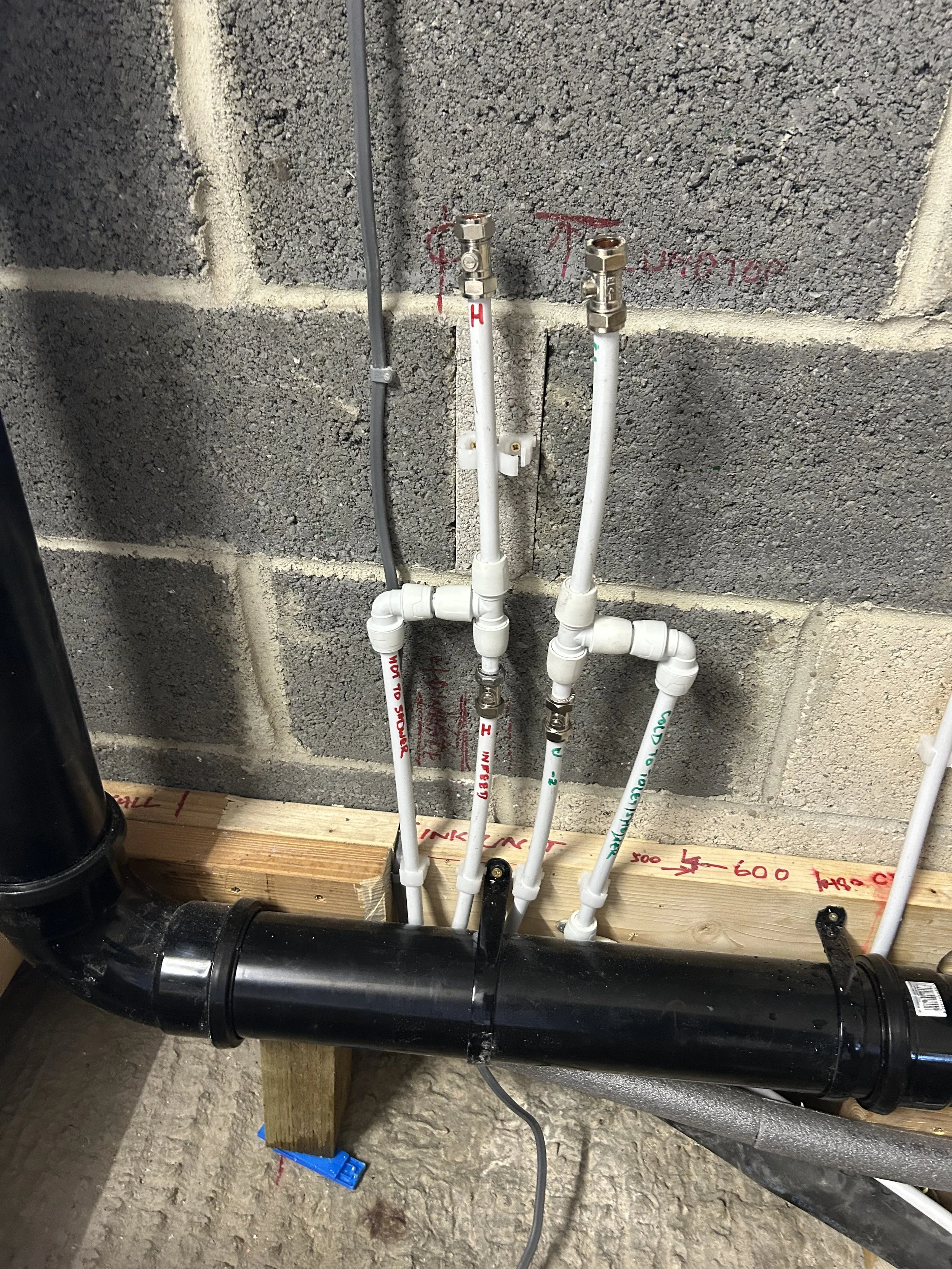 PVC pipes and fittings connected to a large black pipe, mounted on a wooden board against a concrete cinderblock wall.