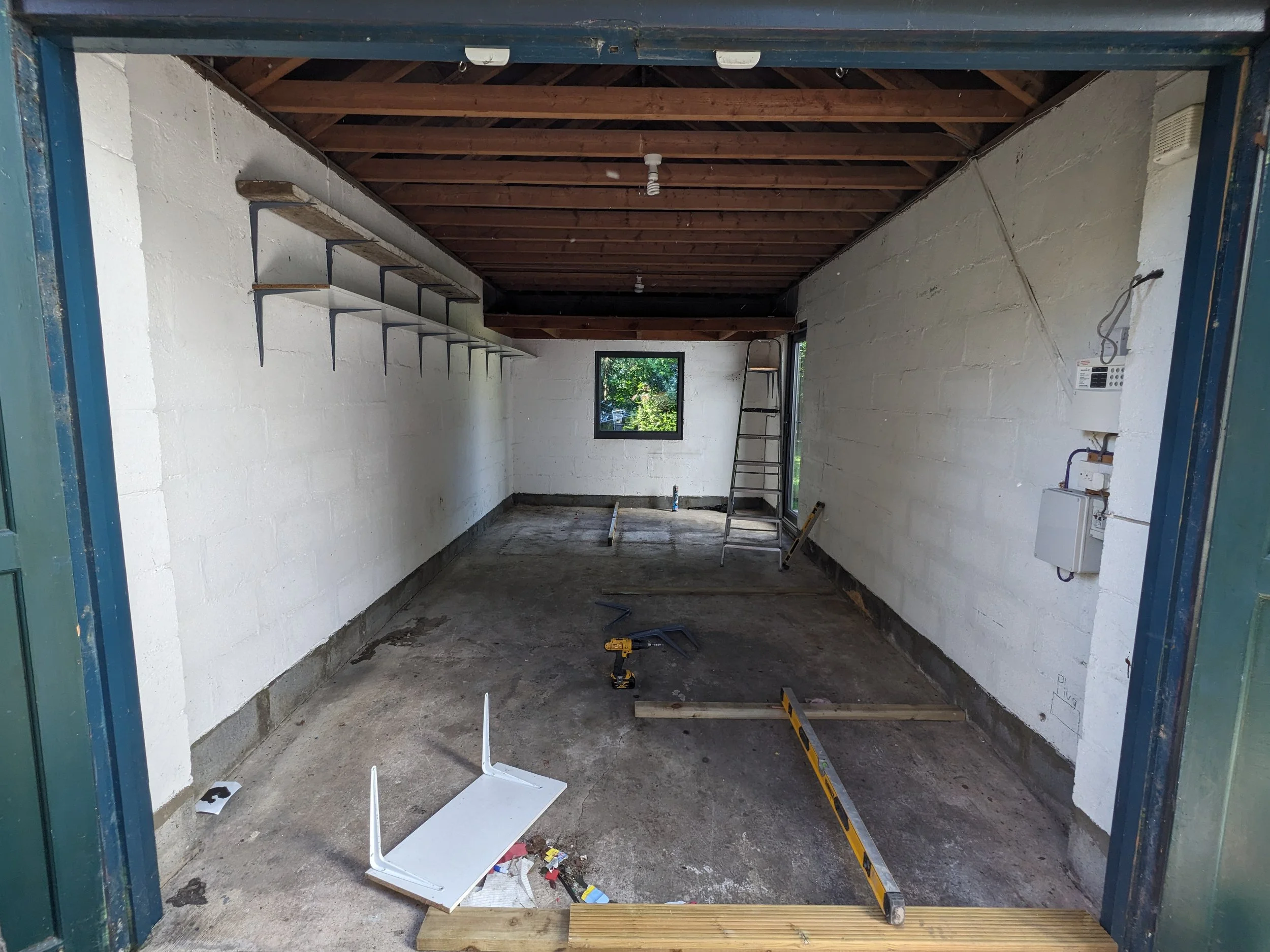 Interior of a garage under construction with tools and supplies on the floor, including a level, a saw, a chair, and a ladder, with white brick walls and a wooden ceiling.
