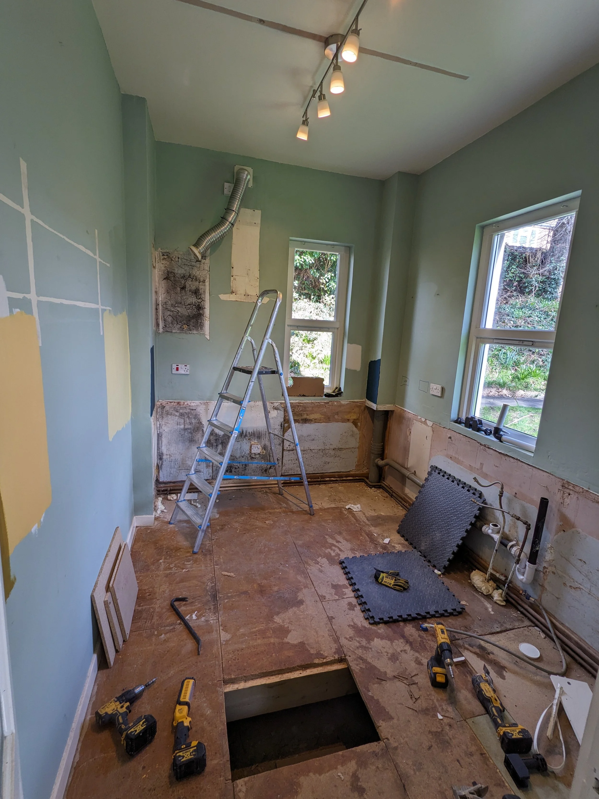 A room under renovation with exposed pipes, a ladder, power tools, and construction materials. The floor is partially removed, revealing a hole, and the walls are partly stripped with patches of paint and drywall. Two windows provide natural light.