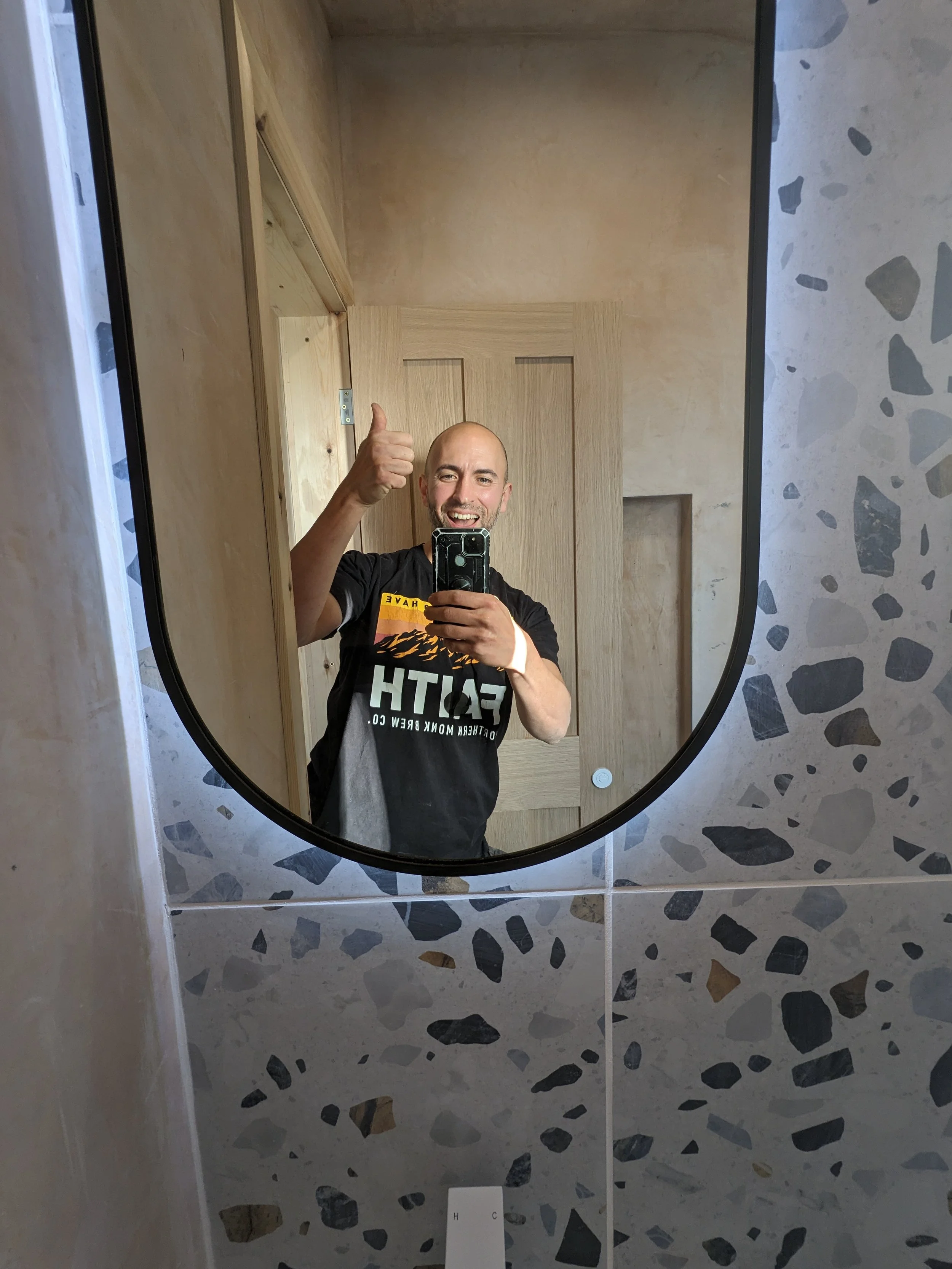 Smiling man taking a mirror selfie in a bathroom, giving a thumbs up.