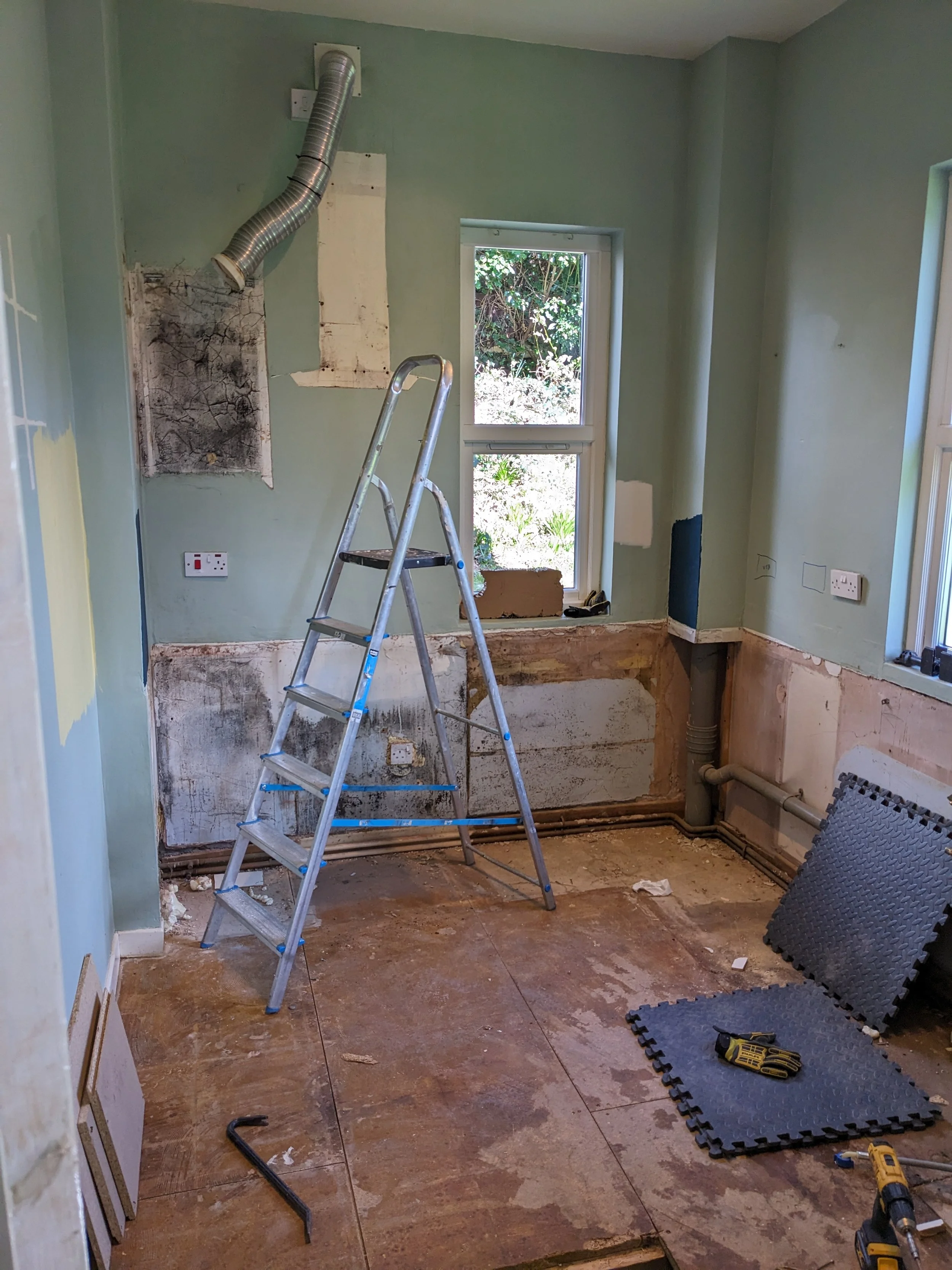 Room under renovation with exposed pipes, electrical outlets, and wall where cabinets or appliances were removed. A ladder, work mats, and tools are on the unfinished floor. Windows let in natural light.