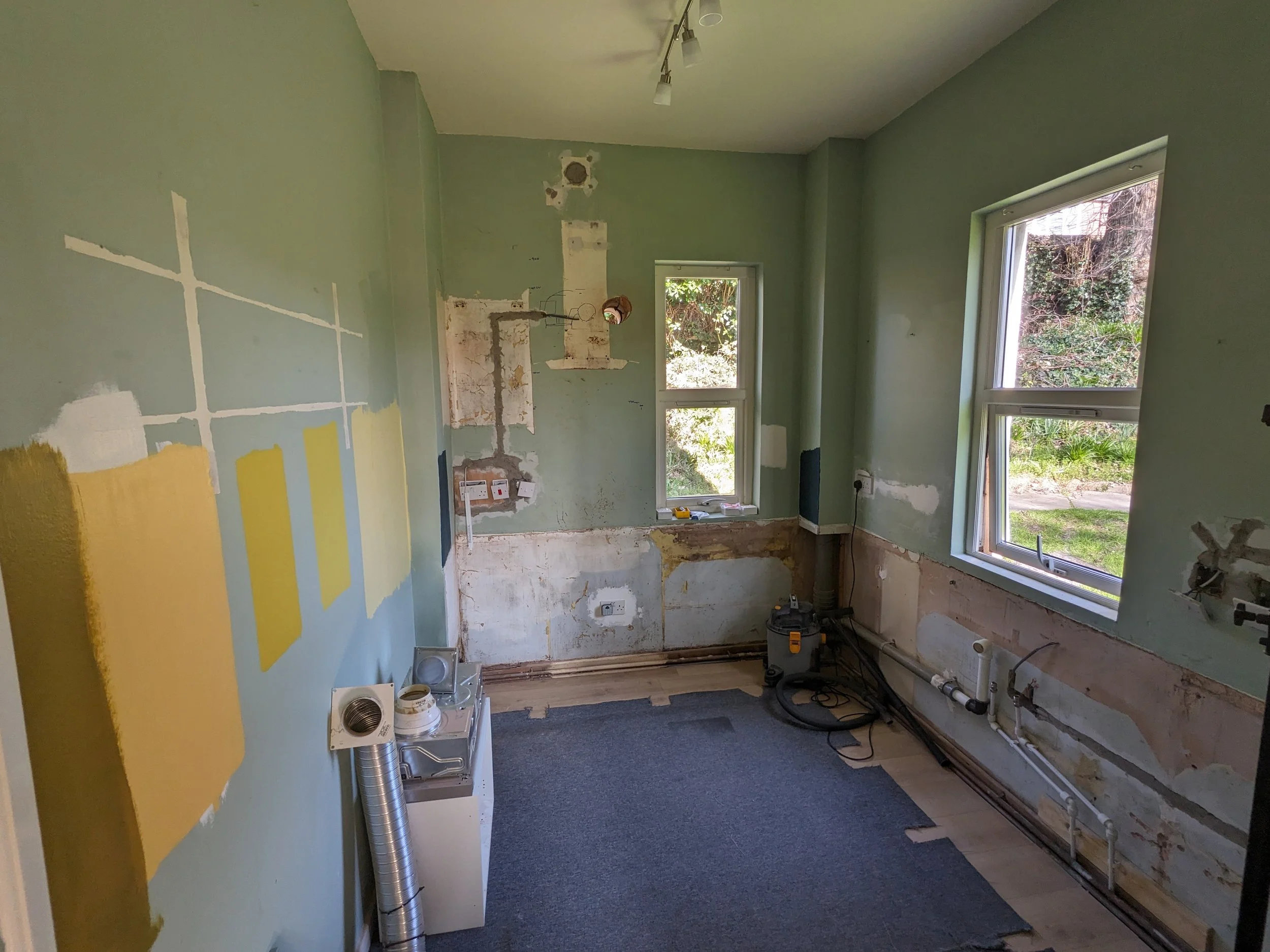 Room under renovation with partially painted walls, exposed pipes and electrical outlets, and construction tools on the floor.