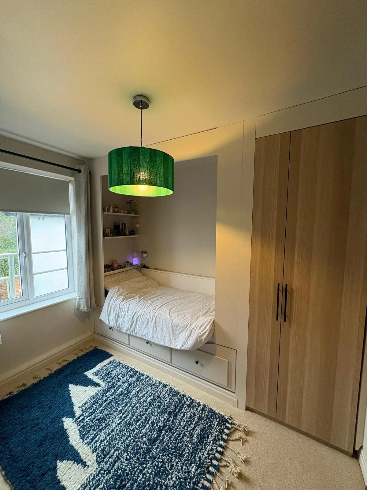 A bedroom with a window, white bed with white bedding, a green ceiling light, a brown wardrobe, and a blue and white patterned rug.