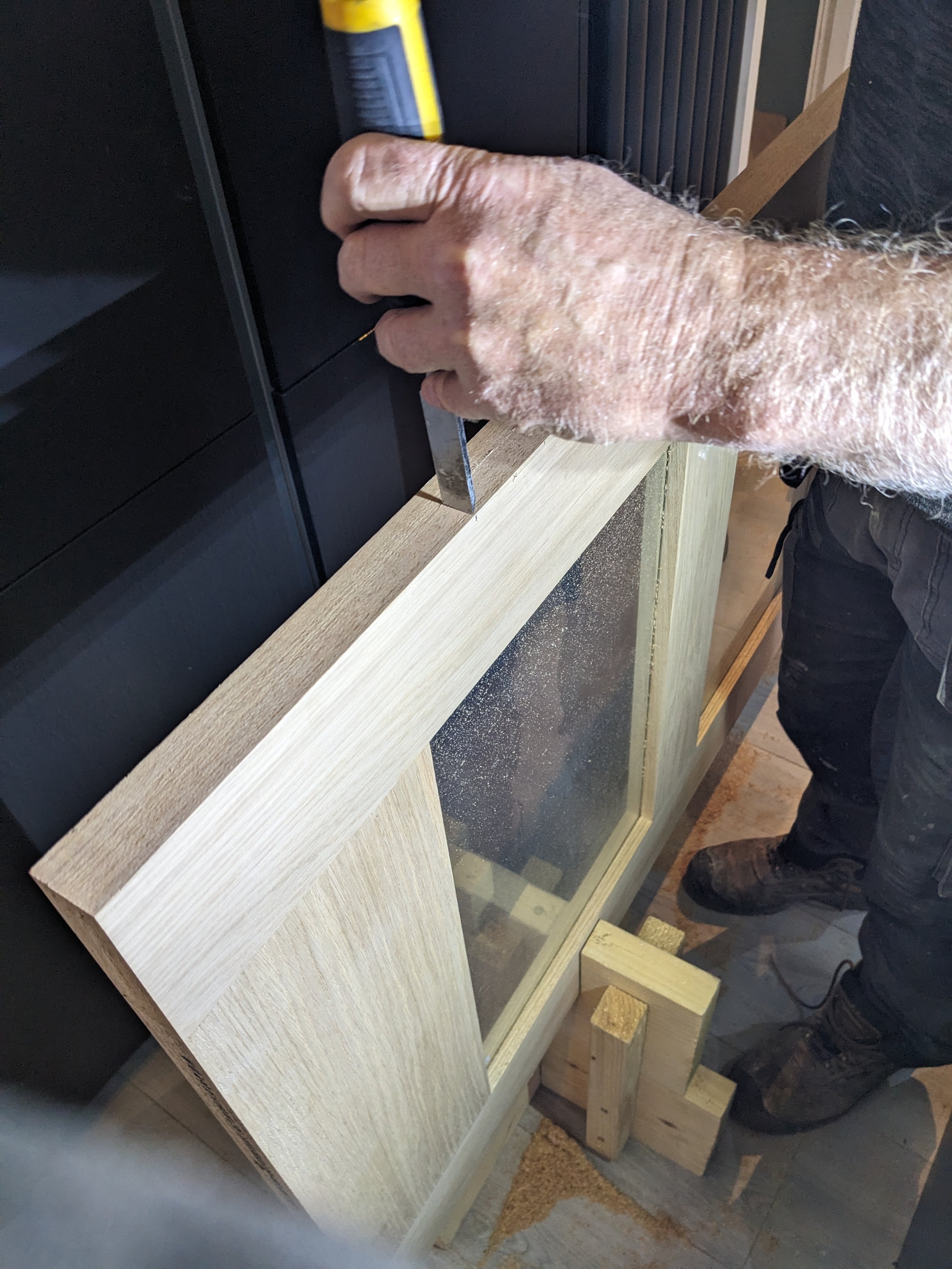 A person using a small chisel or spatula to work on a light-colored wooden frame or cabinet, with black cabinets in the background.