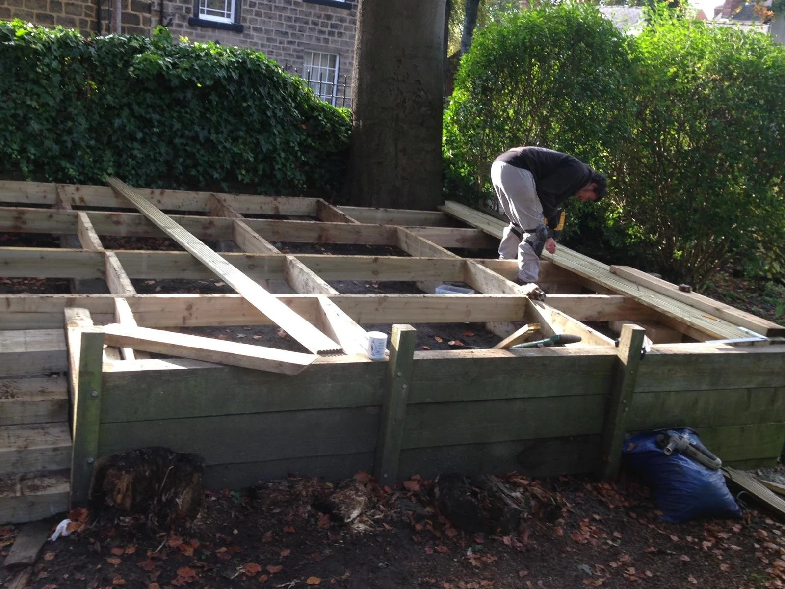 A person working on a wooden deck construction outdoors during daytime, surrounded by trees and bushes.