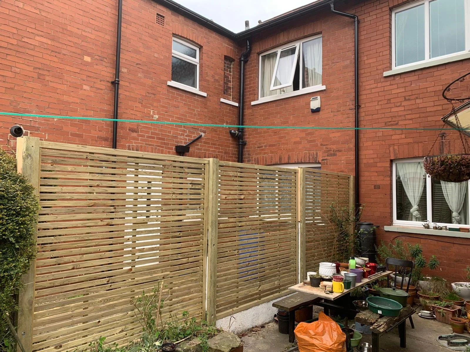 Backyard enclosed by wooden fence with potted plants, gardening supplies on a table, and a brick building with windows, some with curtains, on a cloudy day.