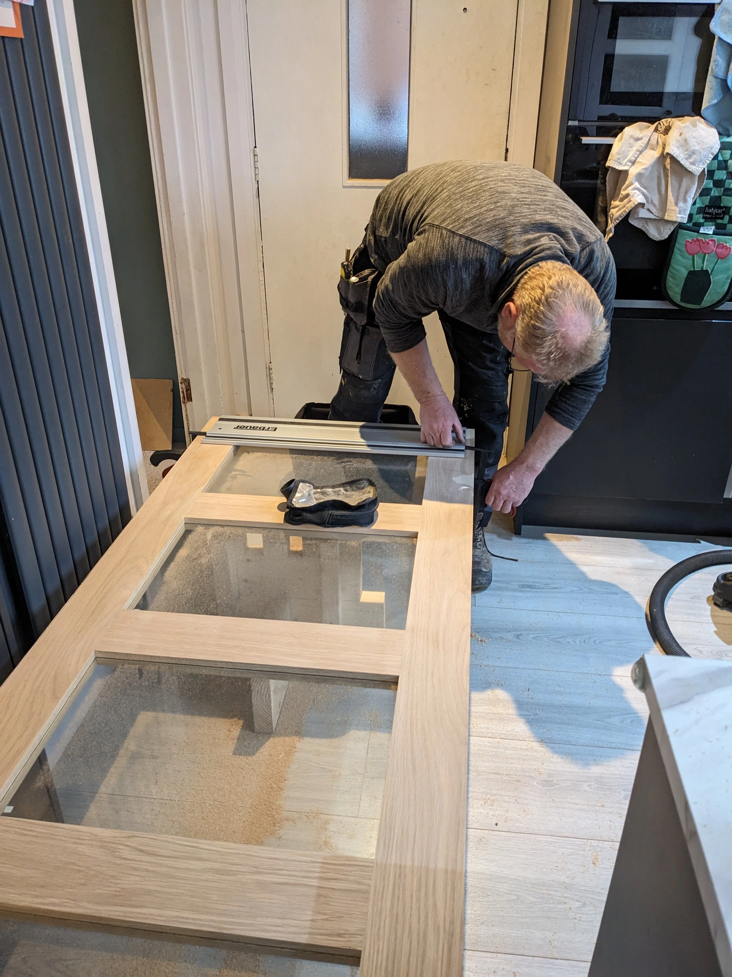 A man working on assembling a wooden piece with glass panels, using a ruler in a room with kitchen and laundry items around.