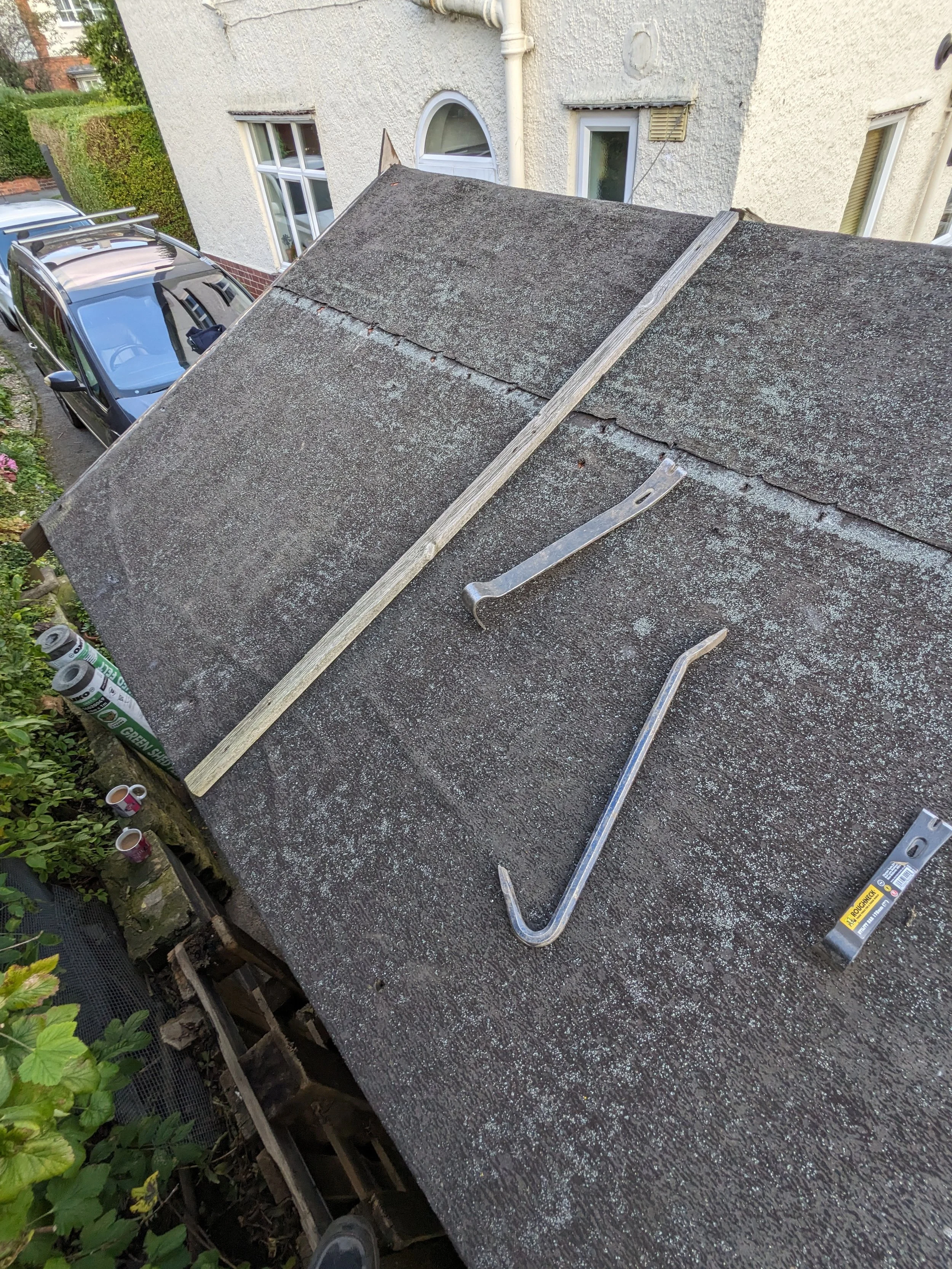 Rooftop with shingles, two crowbars, a wooden plank, and a steel bracket, with a white house and parked cars in the background.