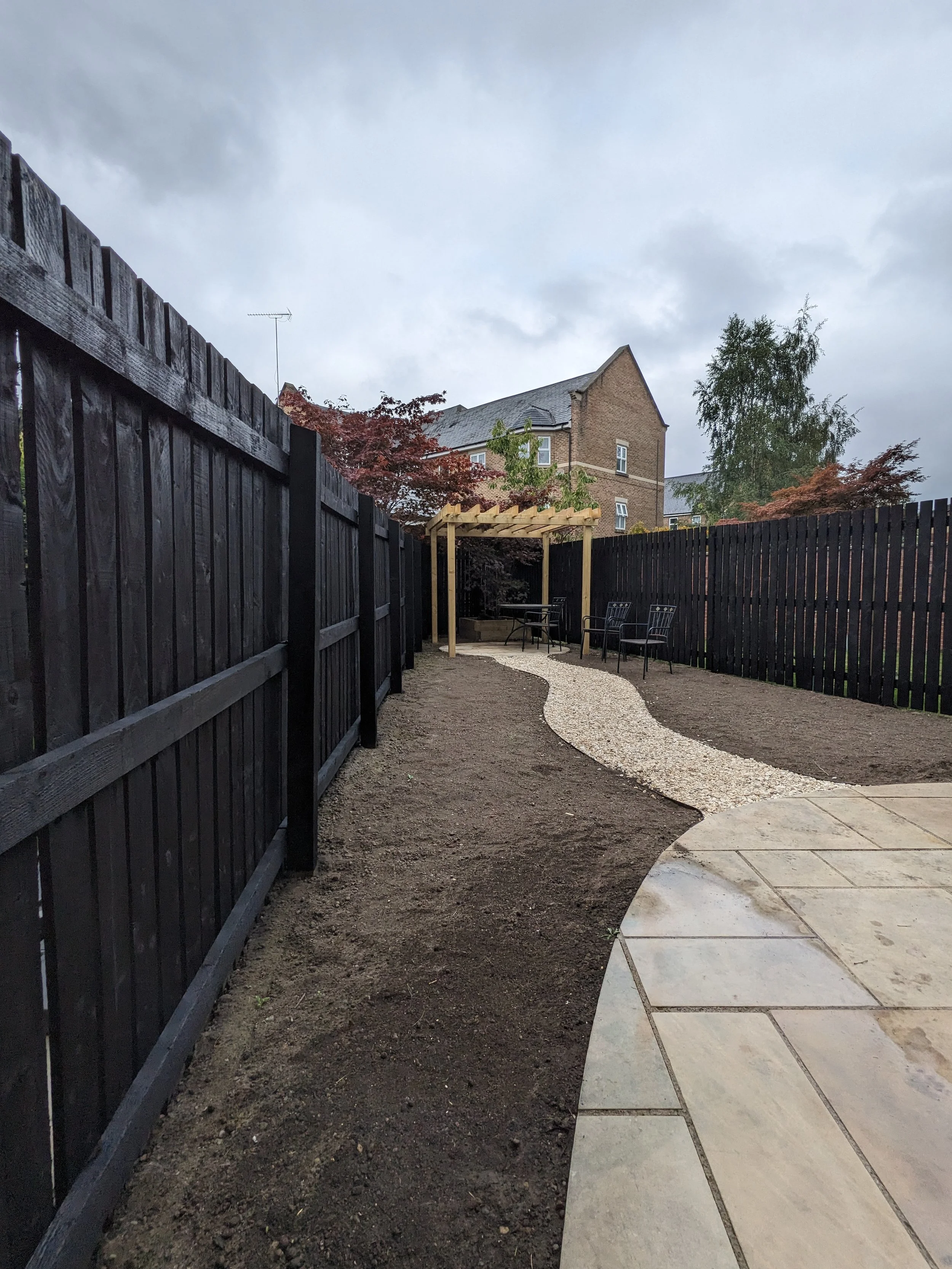 A backyard with a curved gravel pathway leading to a wooden pergola and outdoor seating, enclosed by black wooden fences on either side, with a house and trees in the background under cloudy skies.