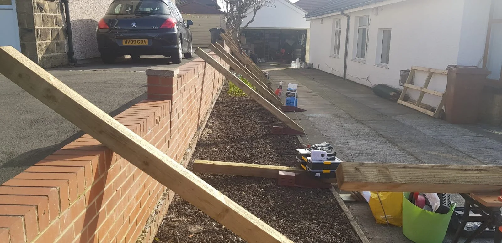 A residential driveway under construction with wooden supports, bricks, tools, and materials spread along the pathway, with a black Peugeot car parked in the background.