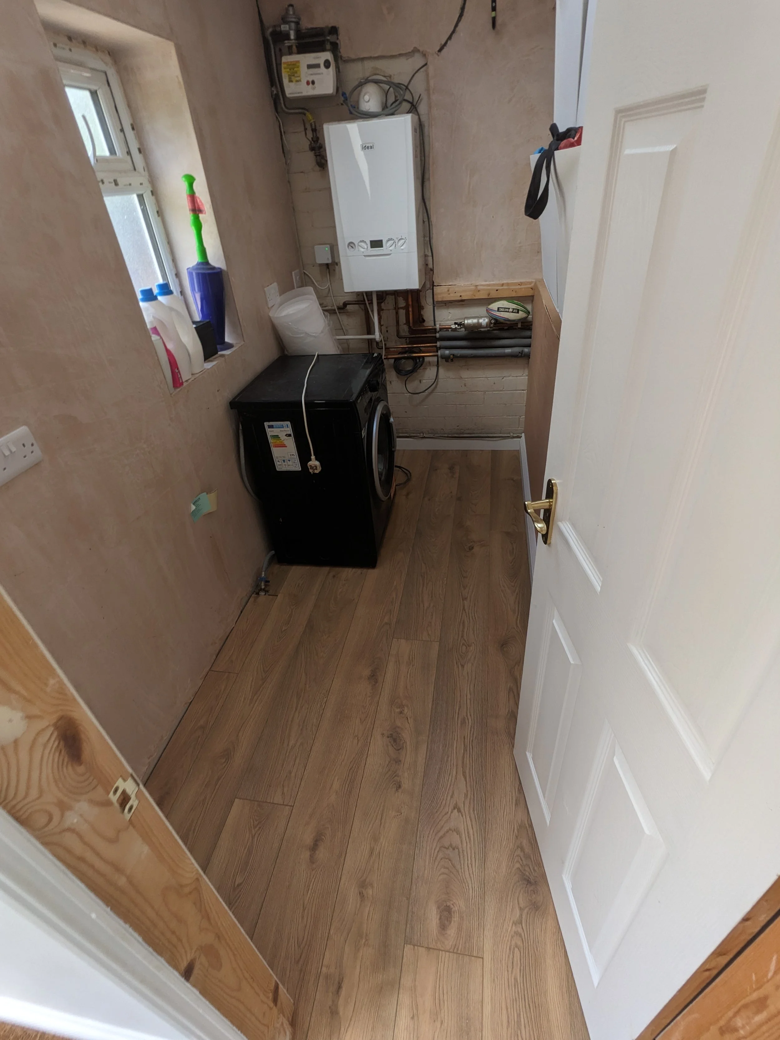 Small utility room with a washing machine, a white wall-mounted boiler, laundry supplies on the window sill, and exposed pipes.