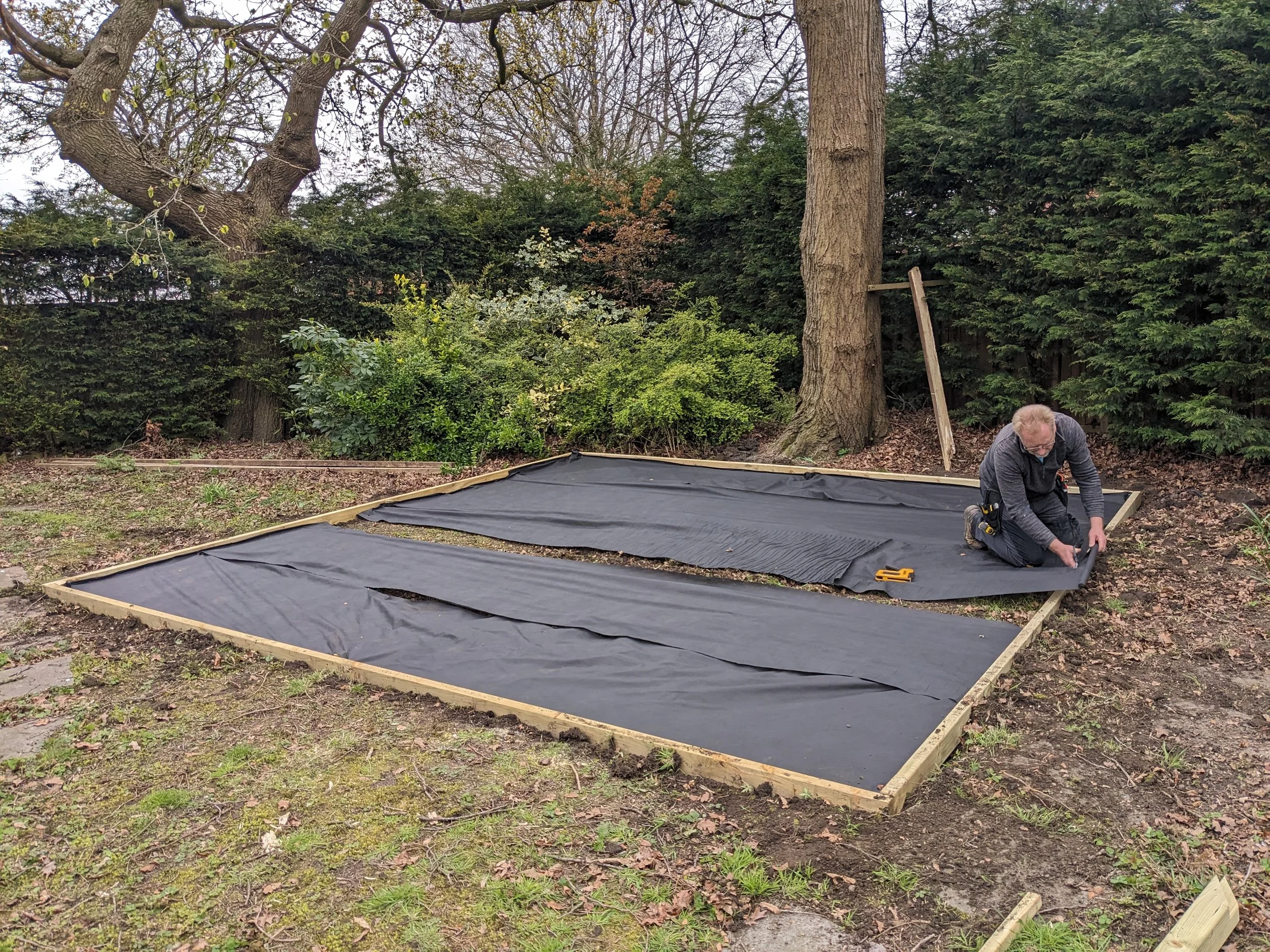 A man installing black pond liner into a wooden frame in a backyard garden with trees and bushes in the background.