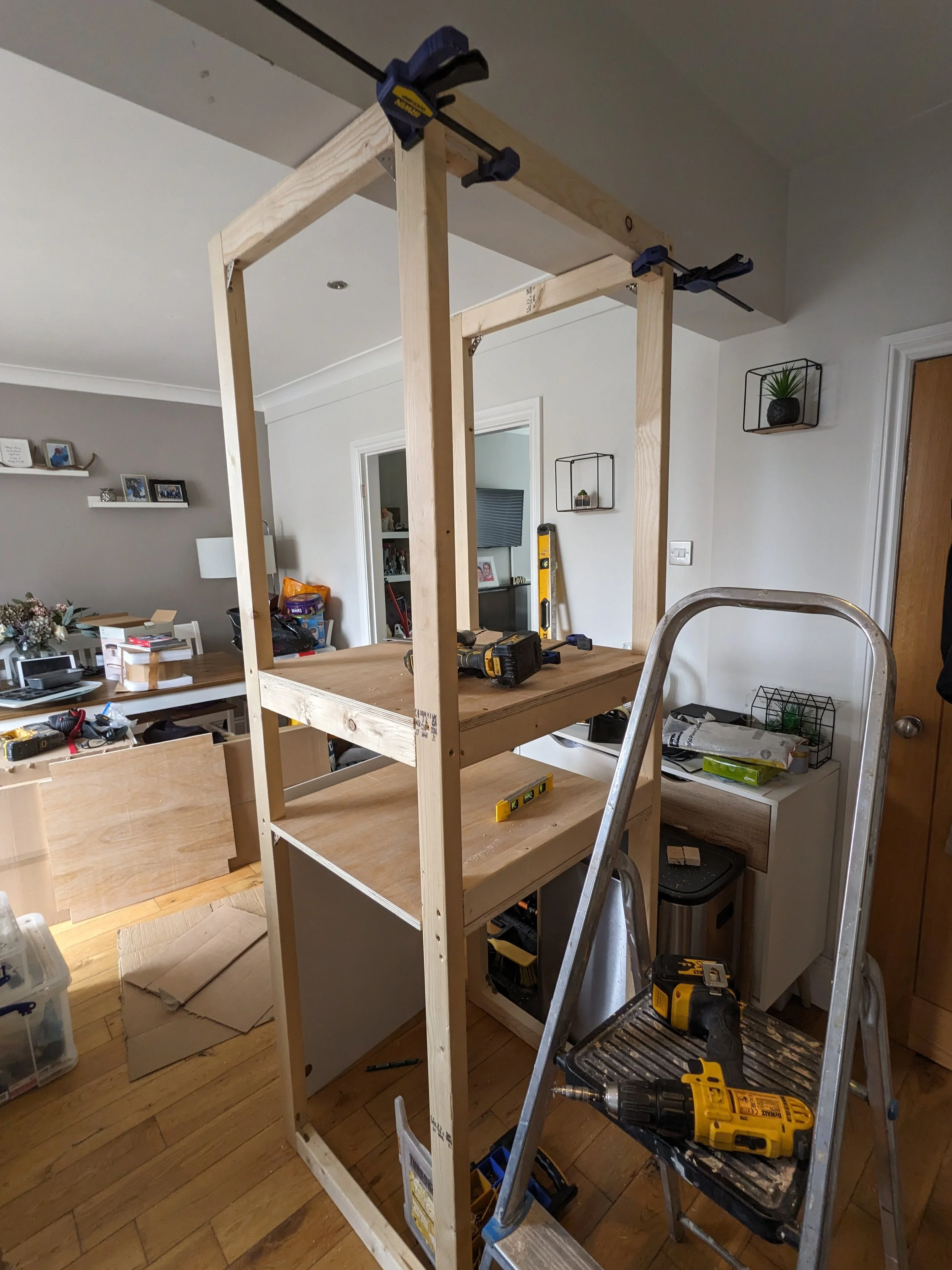 A wooden frame structure under construction in a home, with various tools such as a cordless drill and a level placed on a stool and the frame, set up in a room with furniture and decorations.