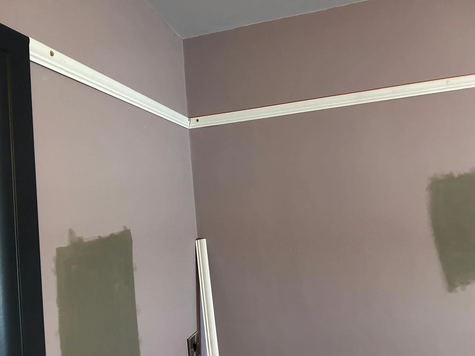 Corner of a room with purple walls and white trim, patches of unpainted wall with patching compound, and a white metal or plastic pipe leaning against the wall.