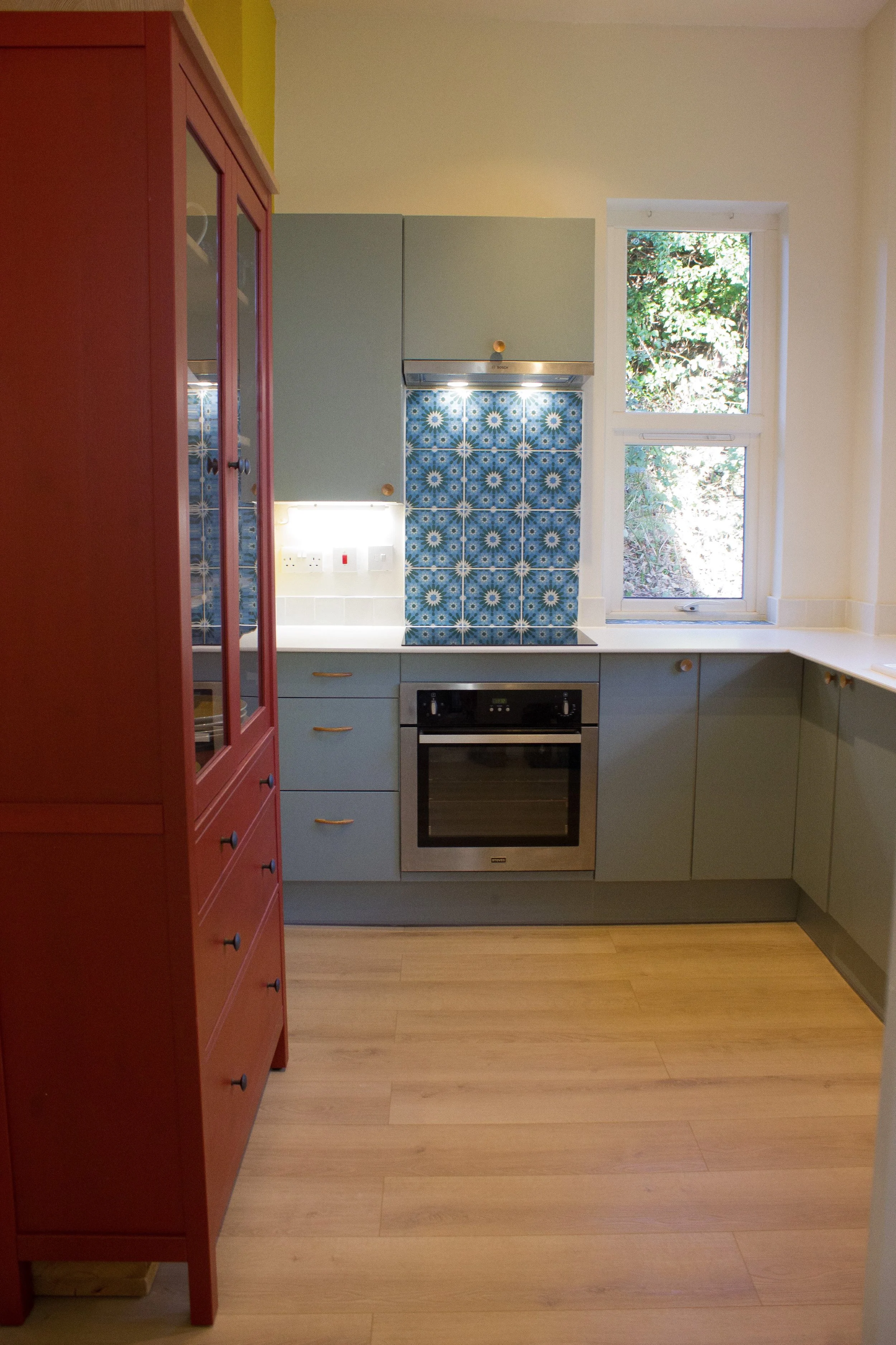 A kitchen with pastel blue cabinets, a blue patterned tiled backsplash, a red storage unit, and a window showing outdoor greenery.
