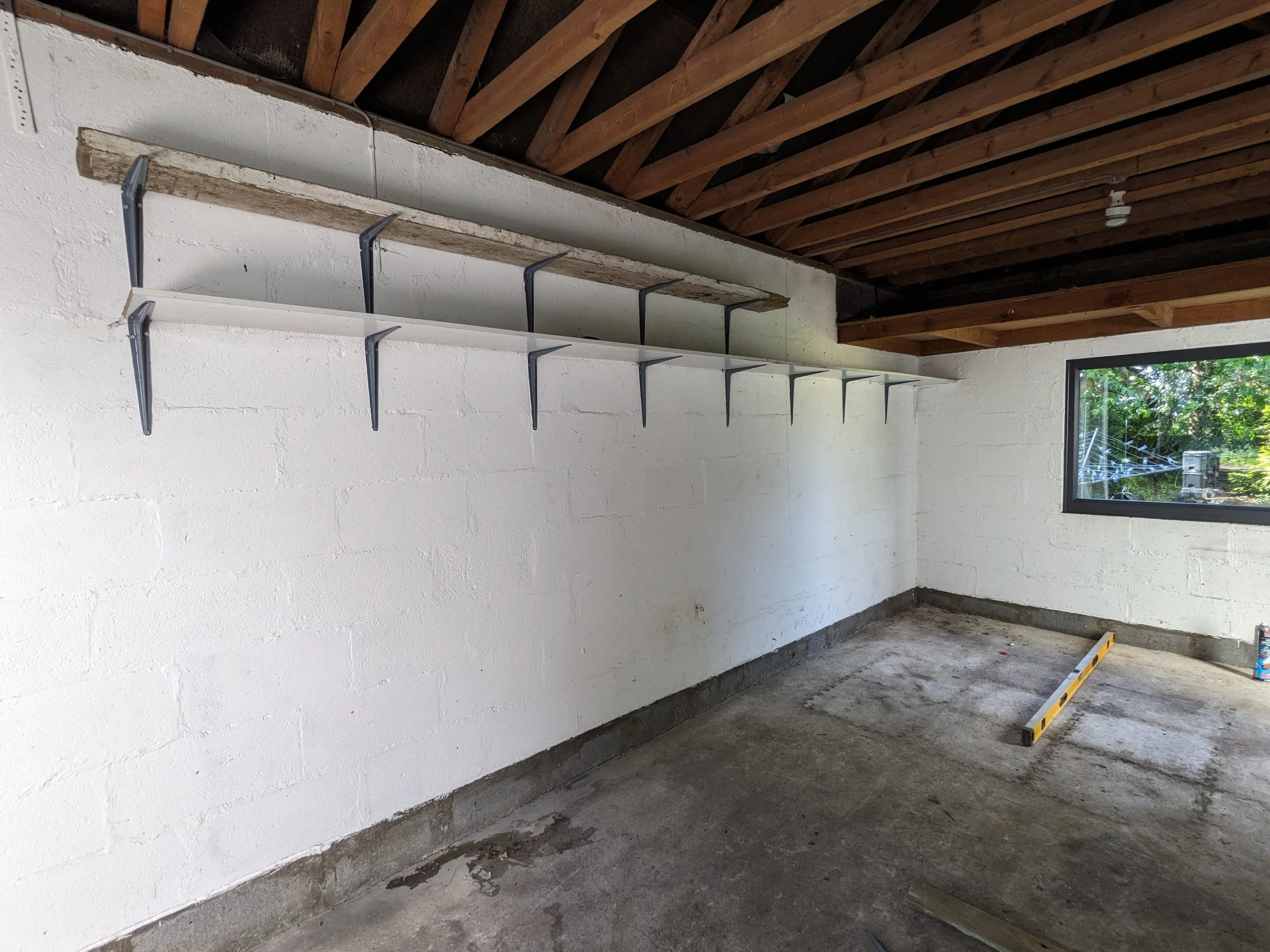 Interior of a garage under construction with white painted brick walls, exposed wooden roof beams, a window showing green trees outside, and a shelf supported by metal brackets mounted on the wall.