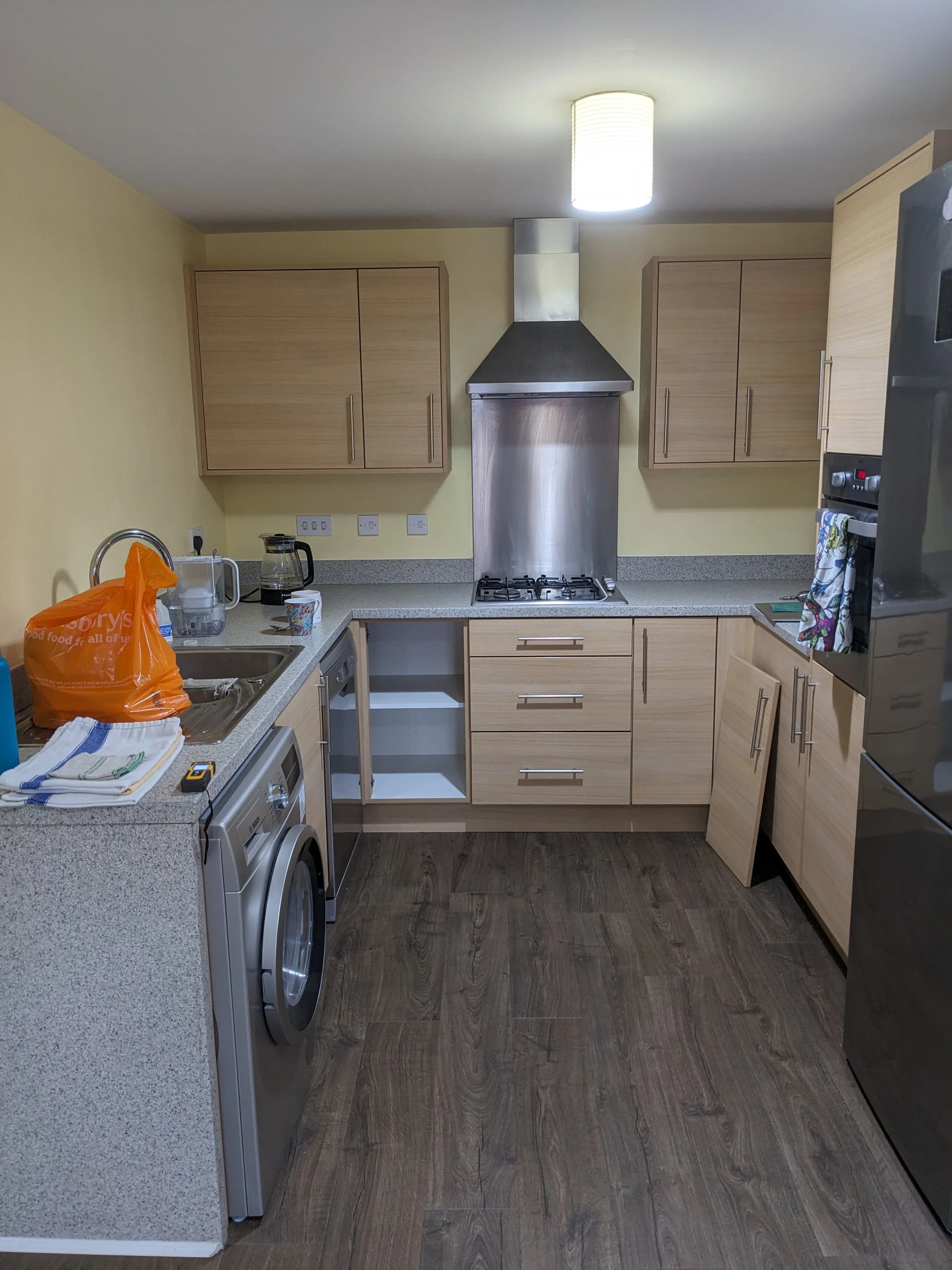 Modern kitchen before photo with wooden cabinets, stainless steel appliances, a washing machine under the counter, and a yellow wall, with items like a plastic bag, coffee maker, and dish towels on the counter.