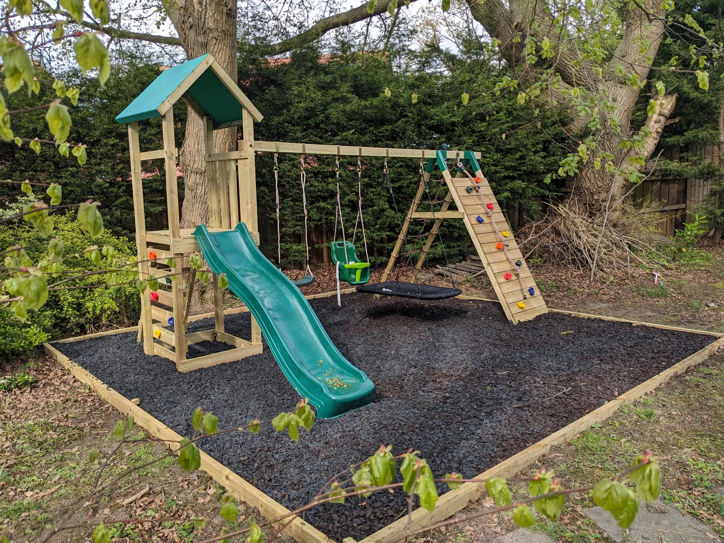 A wooden outdoor playground set with a slide, swings, and a climbing wall, situated on a black gravel surface with trees and bushes in the background.