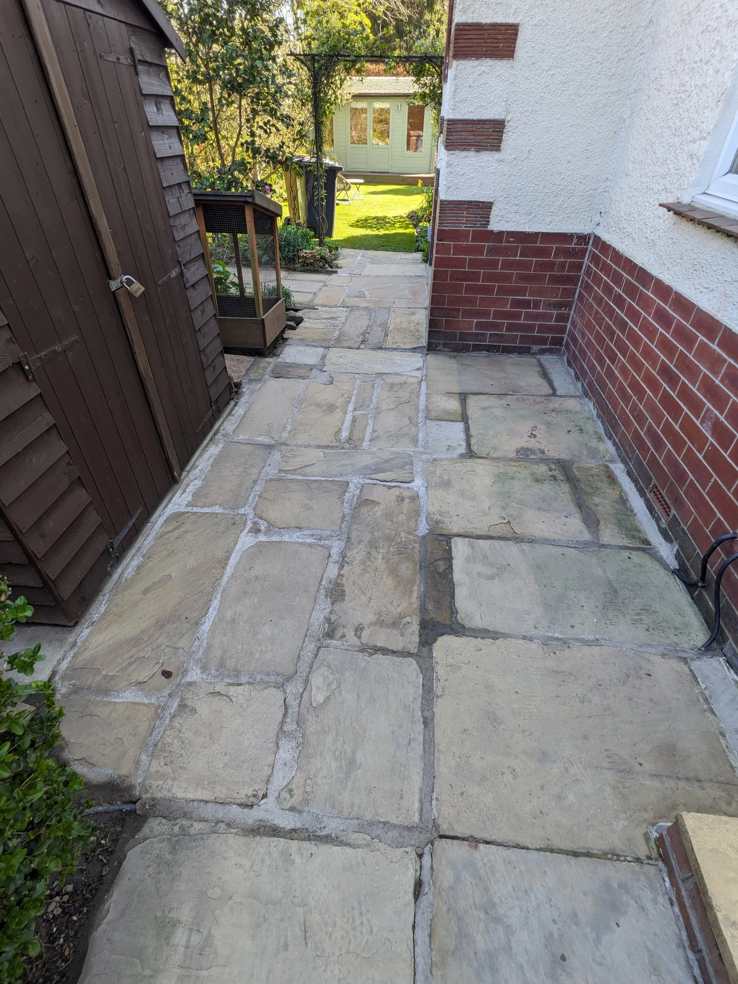 A stone paved pathway leading to a garden with green grass and a small shed in the background, flanked by a dark wooden shed on the left and a white house with red brick accents on the right.