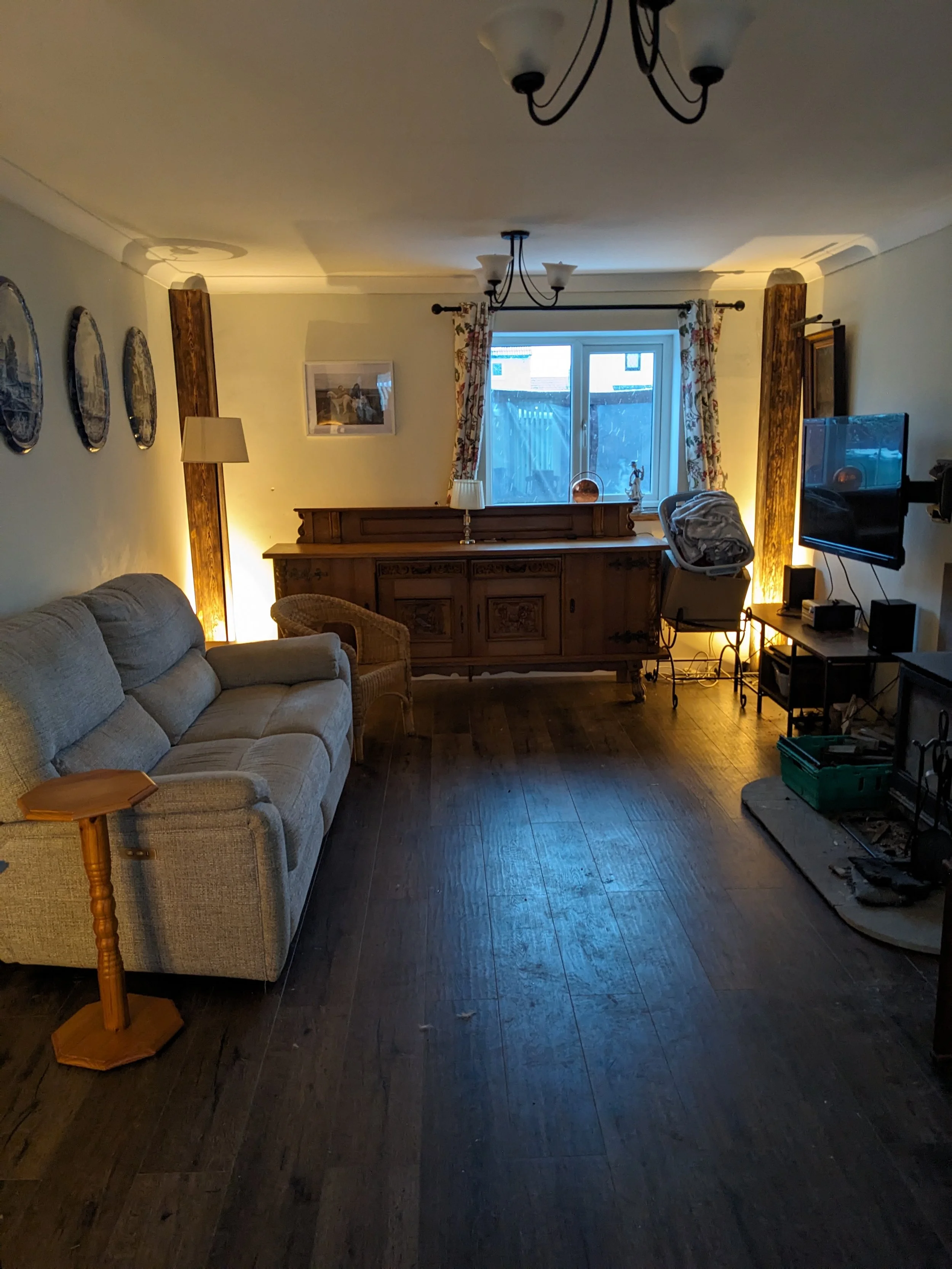 Refurbished living room with beige sofa, wooden floor, window with floral curtains, and television on the right wall.