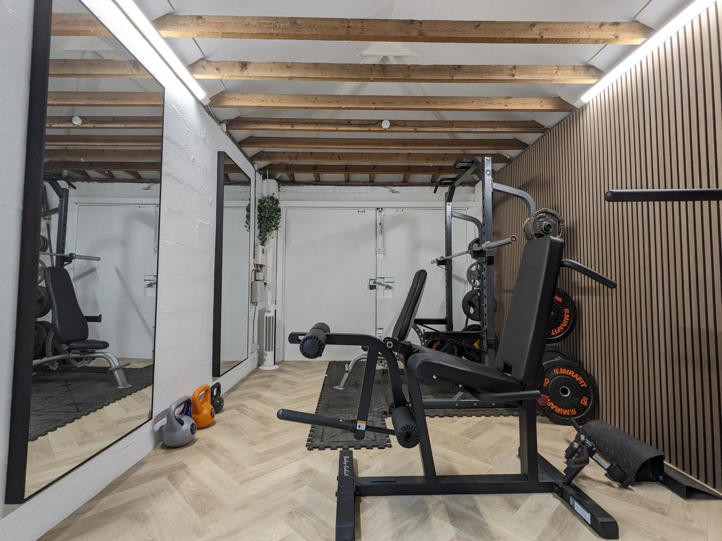 Home gym with weight bench, dumbbells, kettlebells, and weight plates in a room with wooden ceiling beams and a large mirror.