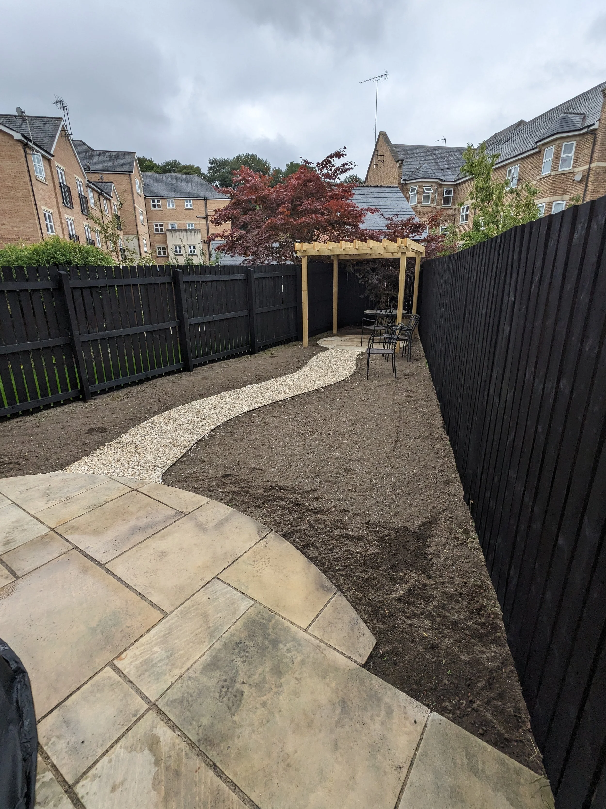 Backyard garden with a newly laid pathway of white pebbles, a small gravel area, and a patio with beige tiles. A black wooden fence surrounds the yard, and a wooden pergola is set up near a red-leaved tree with outdoor furniture underneath.