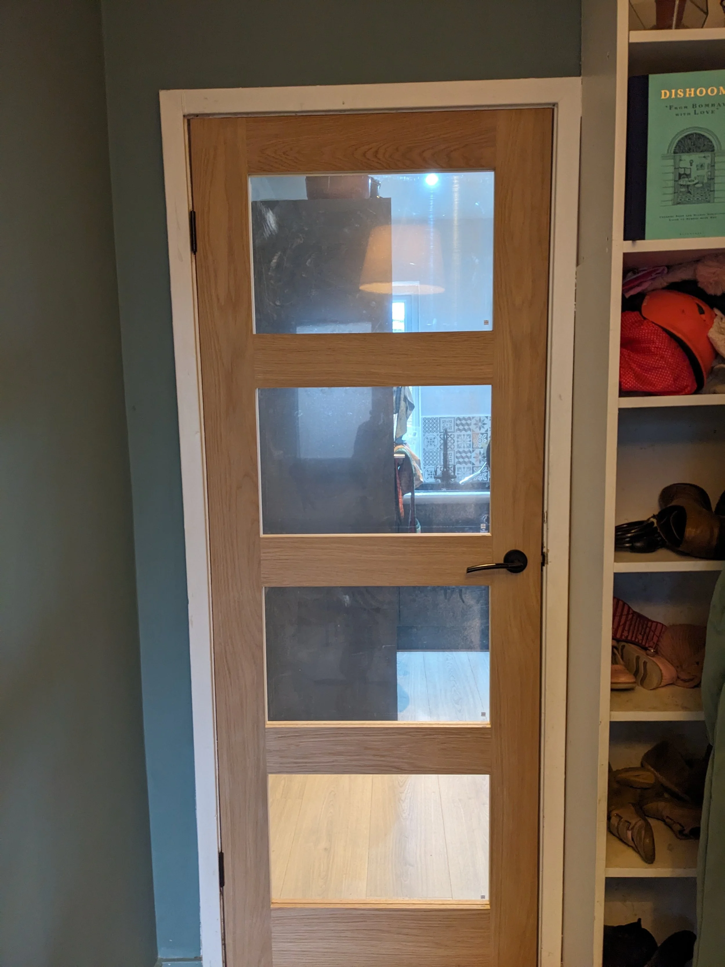 A wooden interior door with four glass panels, partly reflecting the room behind, and a black handle. To the right, a white shelf filled with clothing and other items.