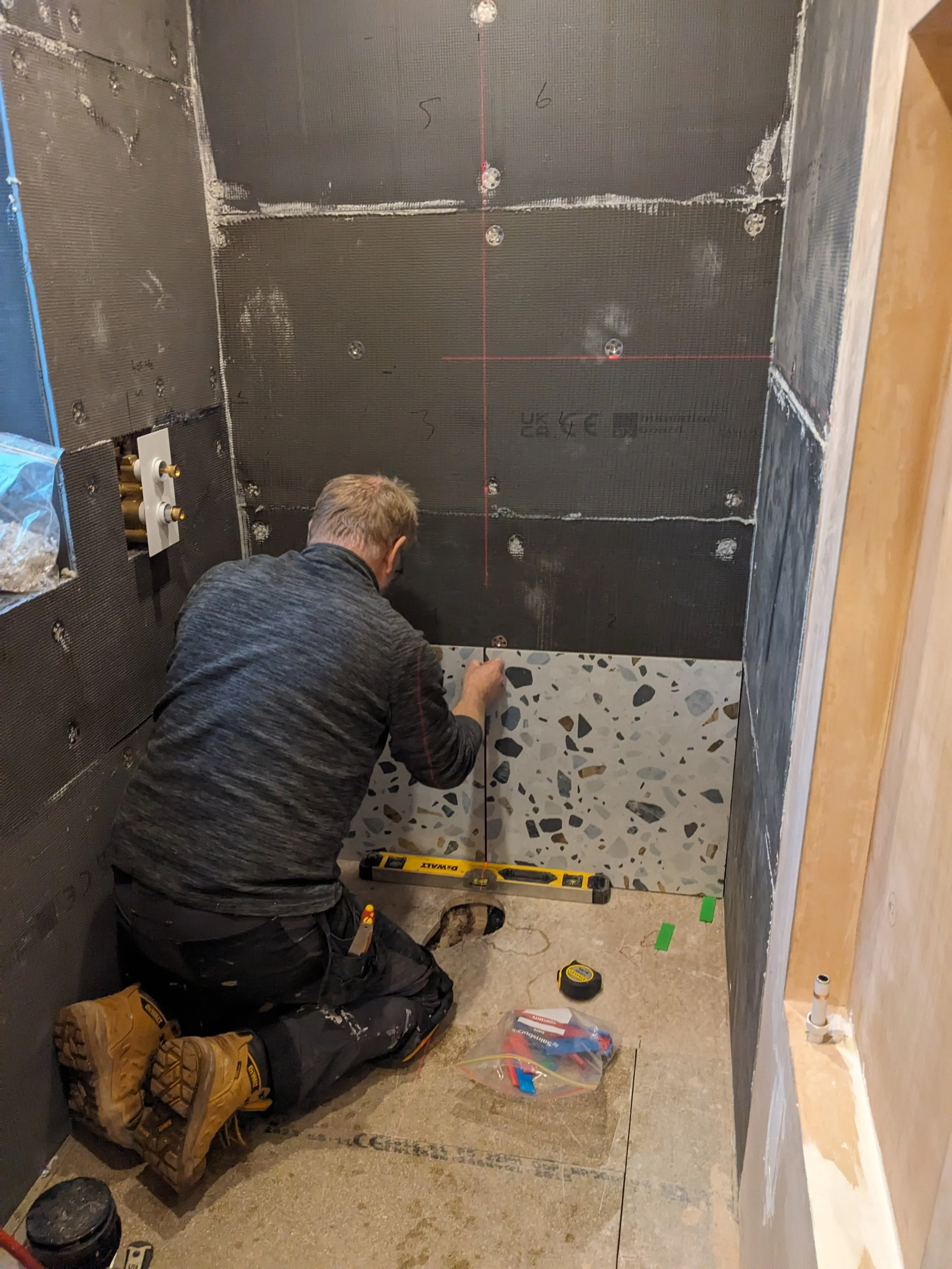 A person kneeling and measuring in a room under construction, with dark insulated walls, a terrazzo tile on the lower wall, and construction tools around.
