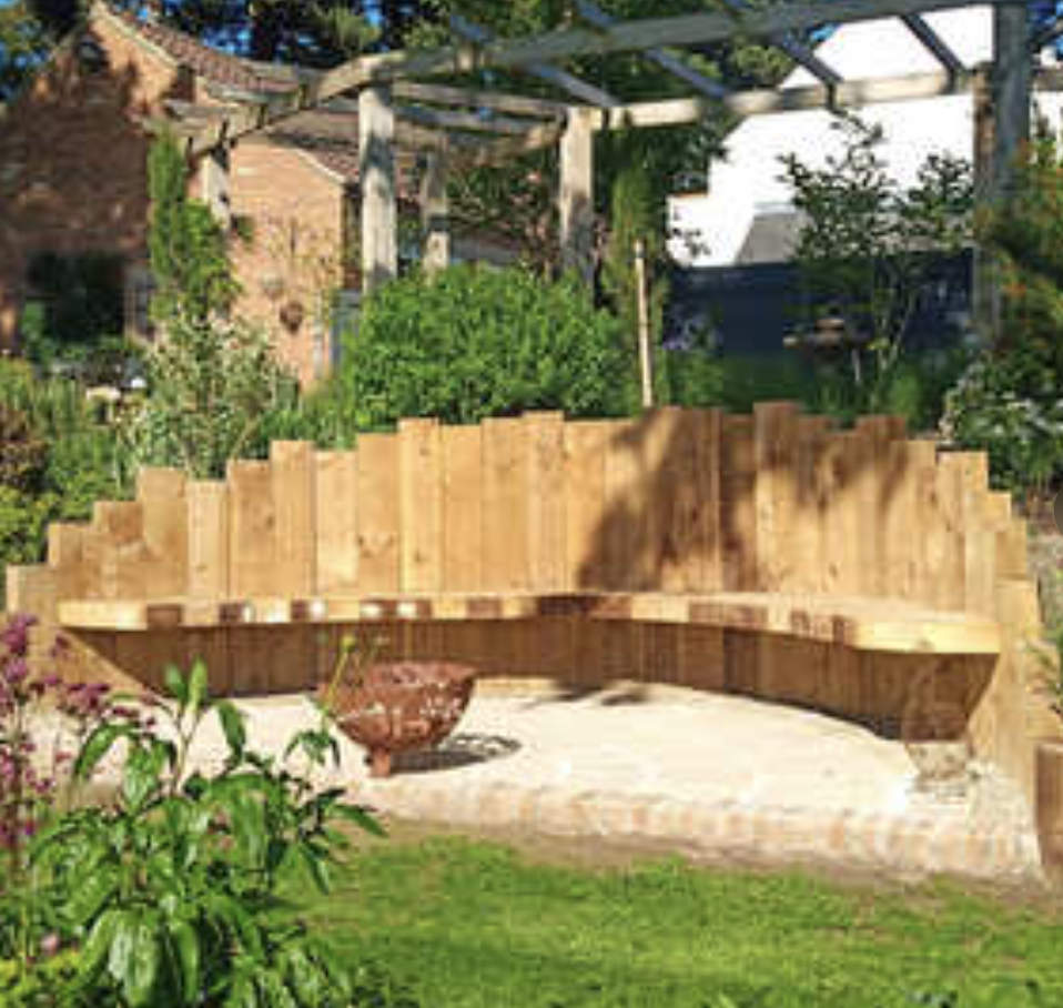 Backyard garden with a curved wooden bench, green plants, trees, and a garden trellis with climbing plants in the background.