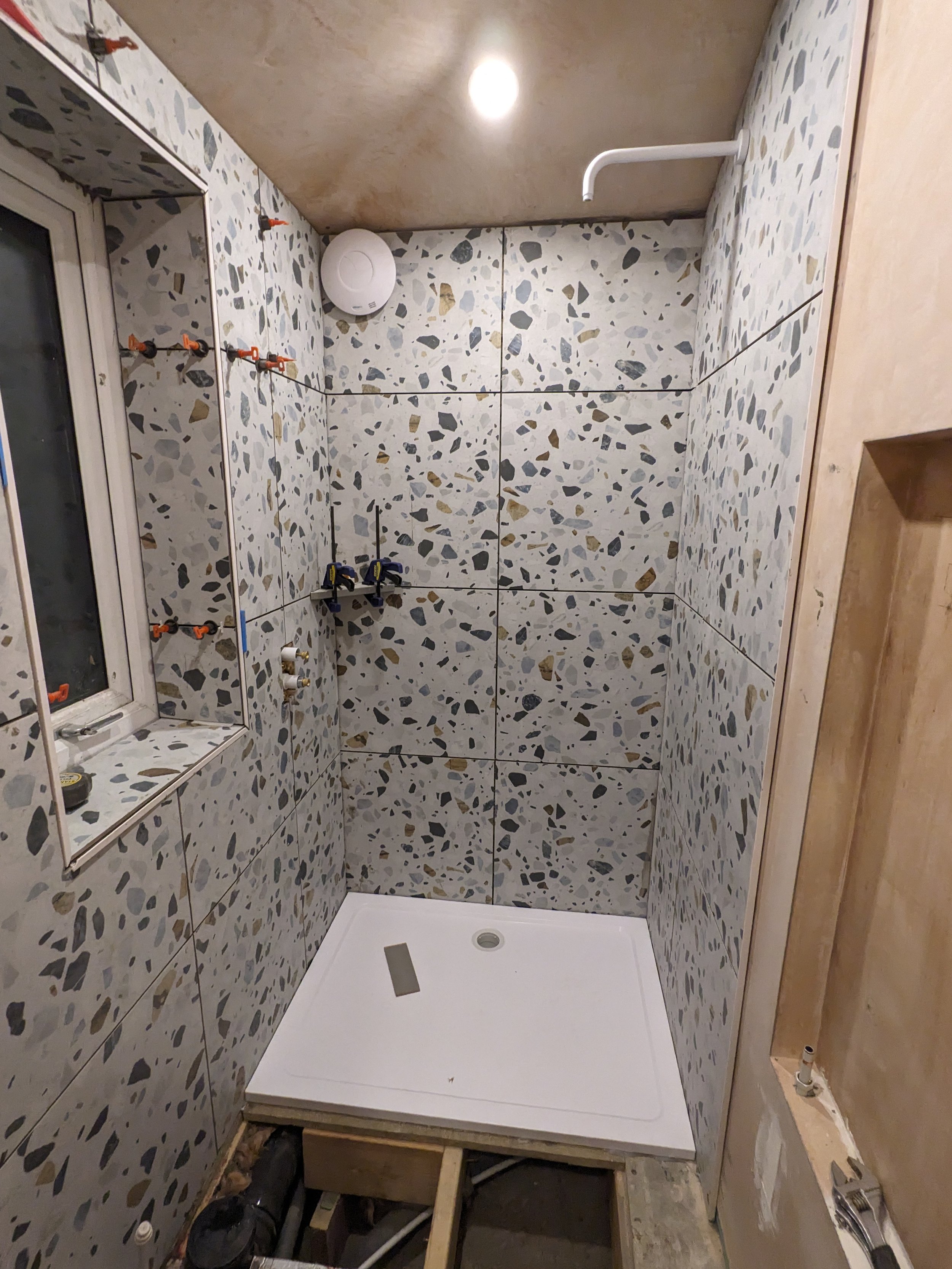 Small bathroom with terrazzo tile walls, white shower base on a wooden platform, construction tools, and pipes for plumbing, with an unfinished wall and a small window.