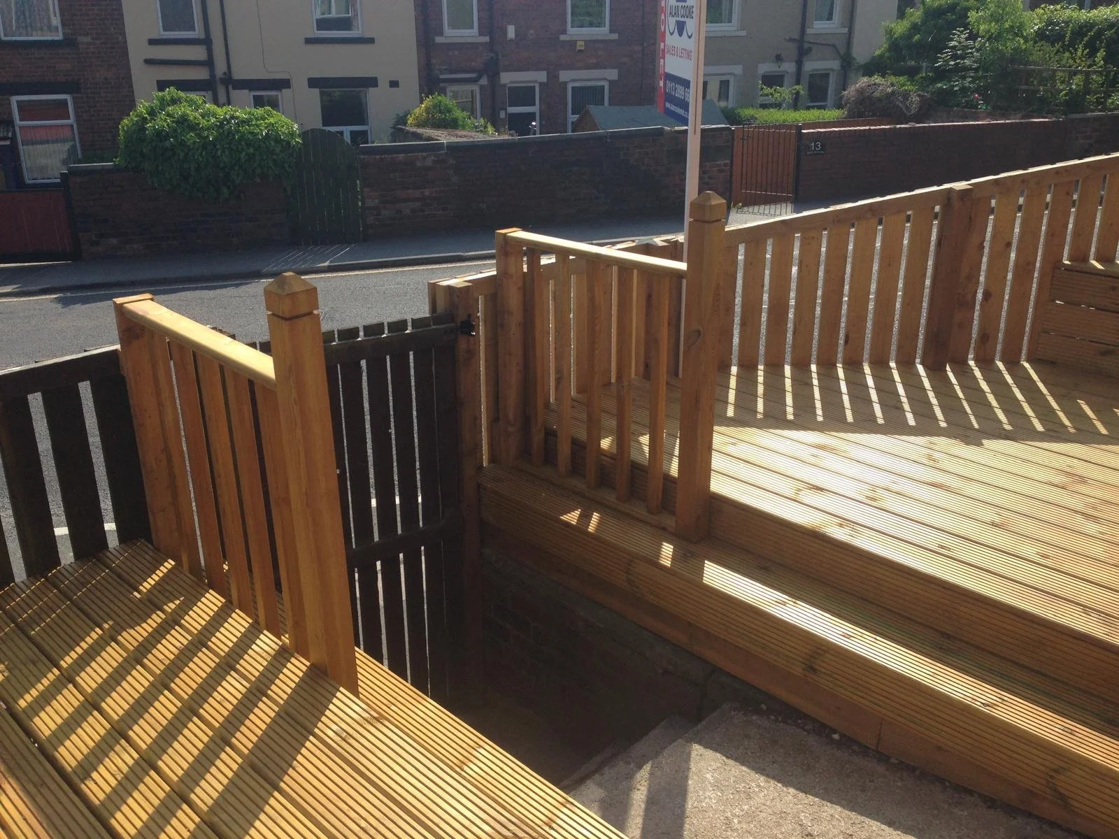 View of a wooden deck with railings, in a residential area, with a street and brick wall in the background, and a painted sign on a pole outside the fence.