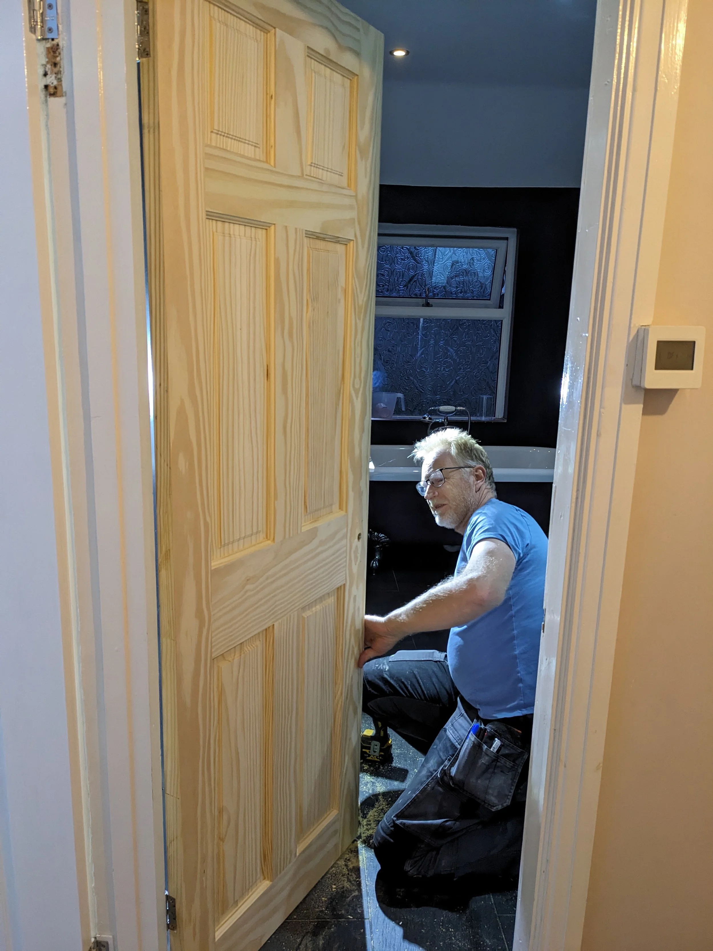 A man installing a wooden door in a room with a window and a bathtub in the background.