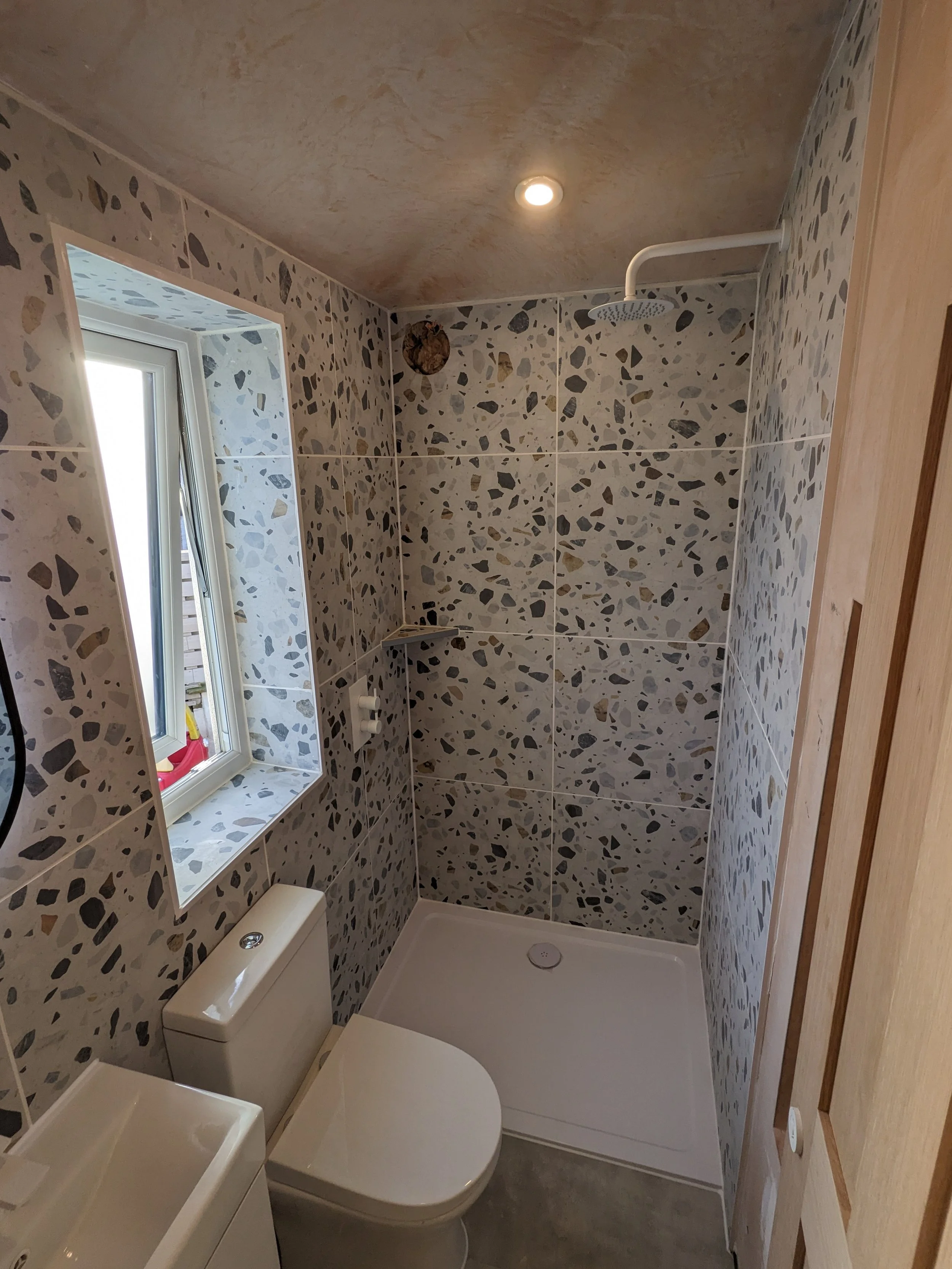 Small bathroom with terrazzo wall tiles, a toilet, a small sink, a window, a shower area with a white square shower tray, and a ceiling with a recessed light.