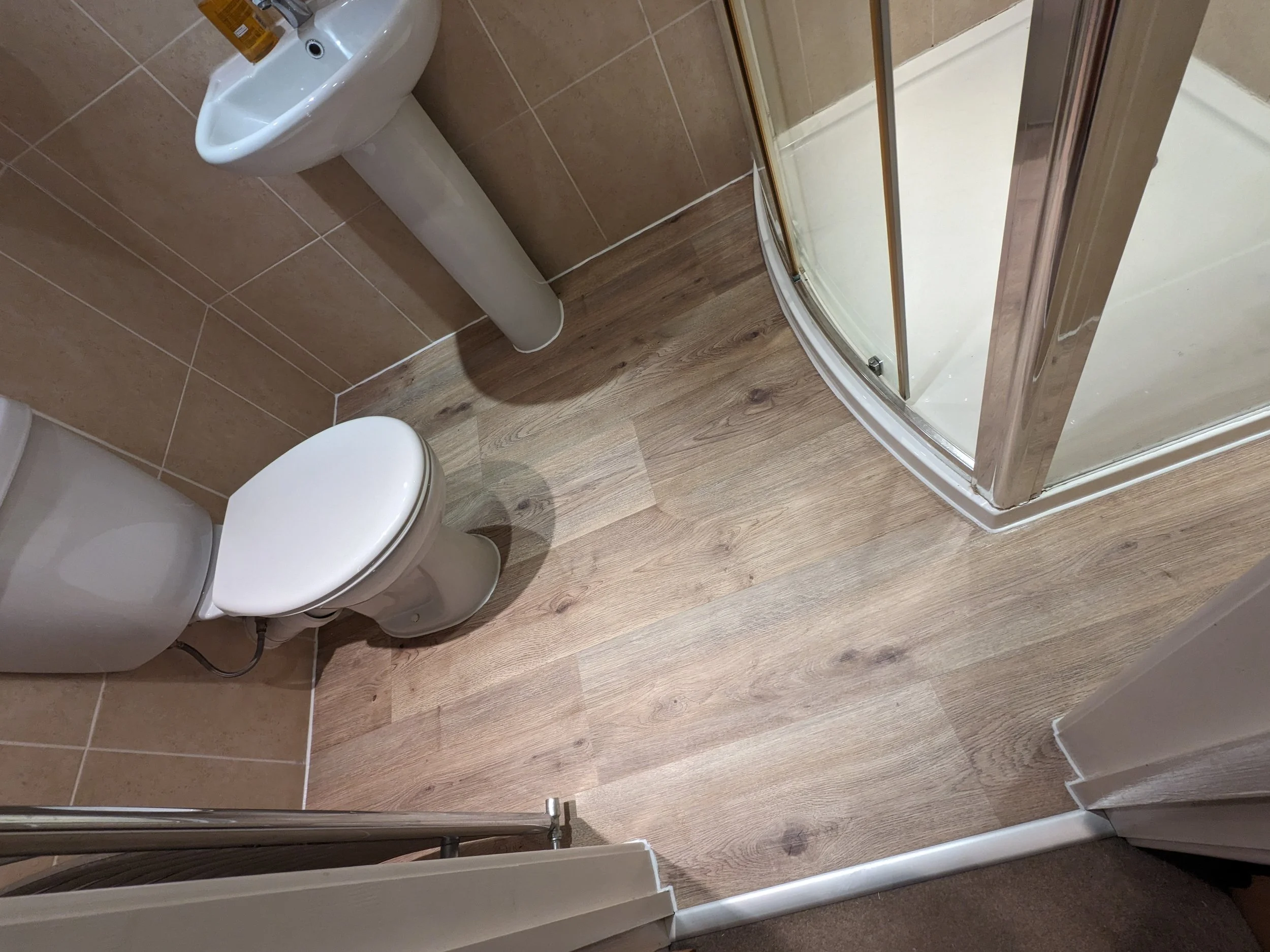 A small bathroom with a toilet, a bidet, a corner shower with a curved glass door, and wood-style flooring with beige tiles on the wall.