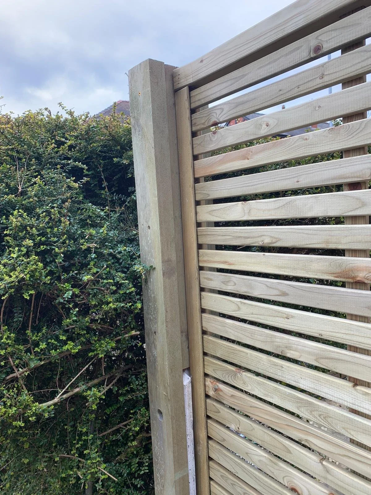 A newly installed wooden fence with horizontal slats next to dense green shrubbery on a cloudy day.
