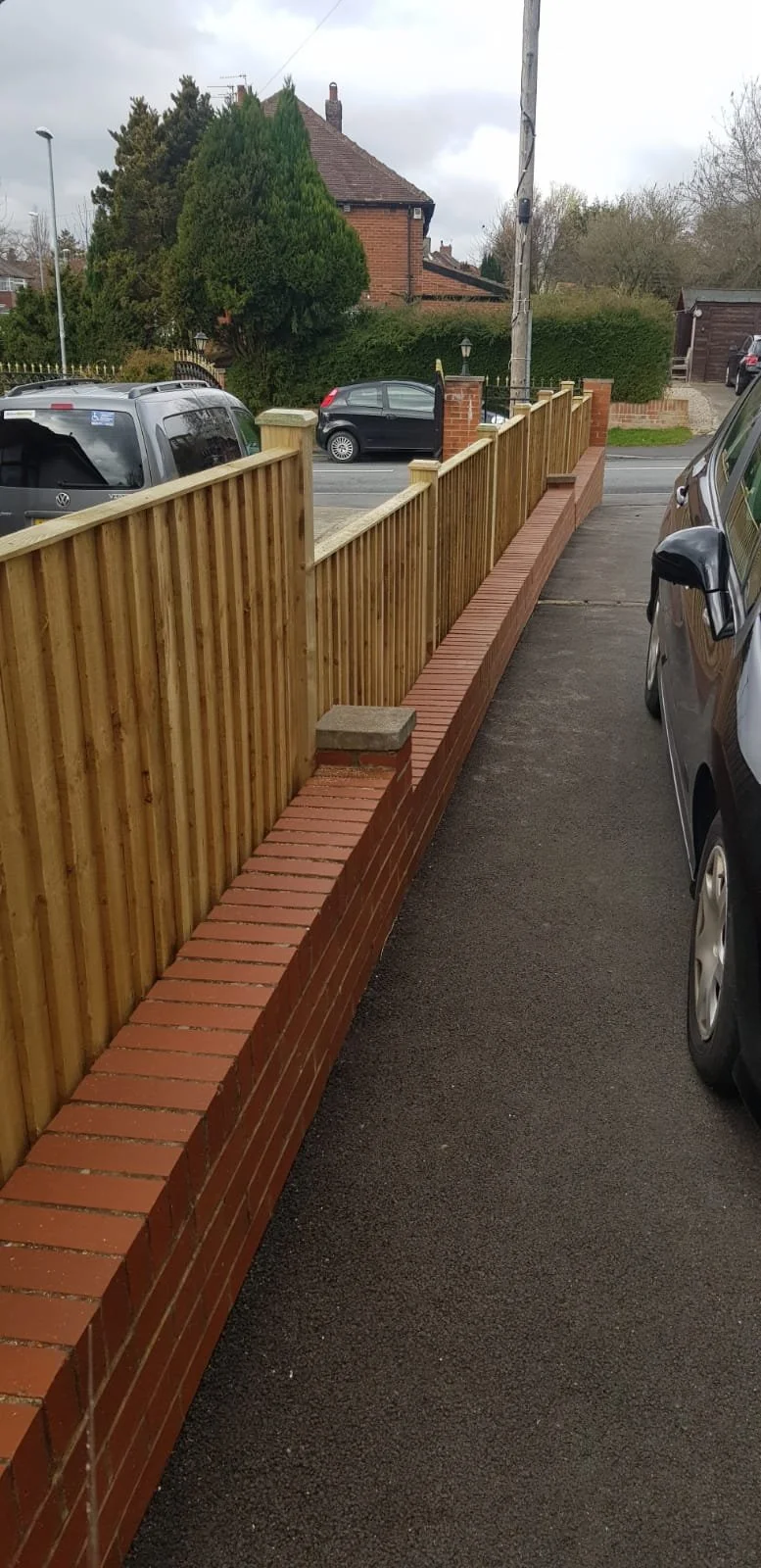 View of a residential street with a newly built wooden fence atop a brick wall separating a driveway from the sidewalk. Parked cars are visible along the street, and a house with trees is in the background under an overcast sky.