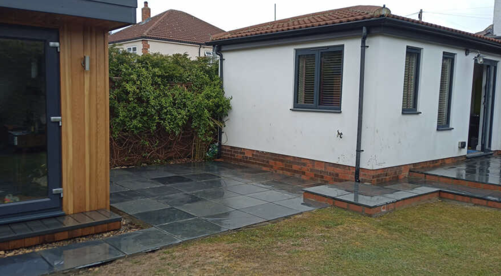 Backyard of a house with black stone paving, a white exterior wall, windows with shutters, green bush, and steps leading to a door.