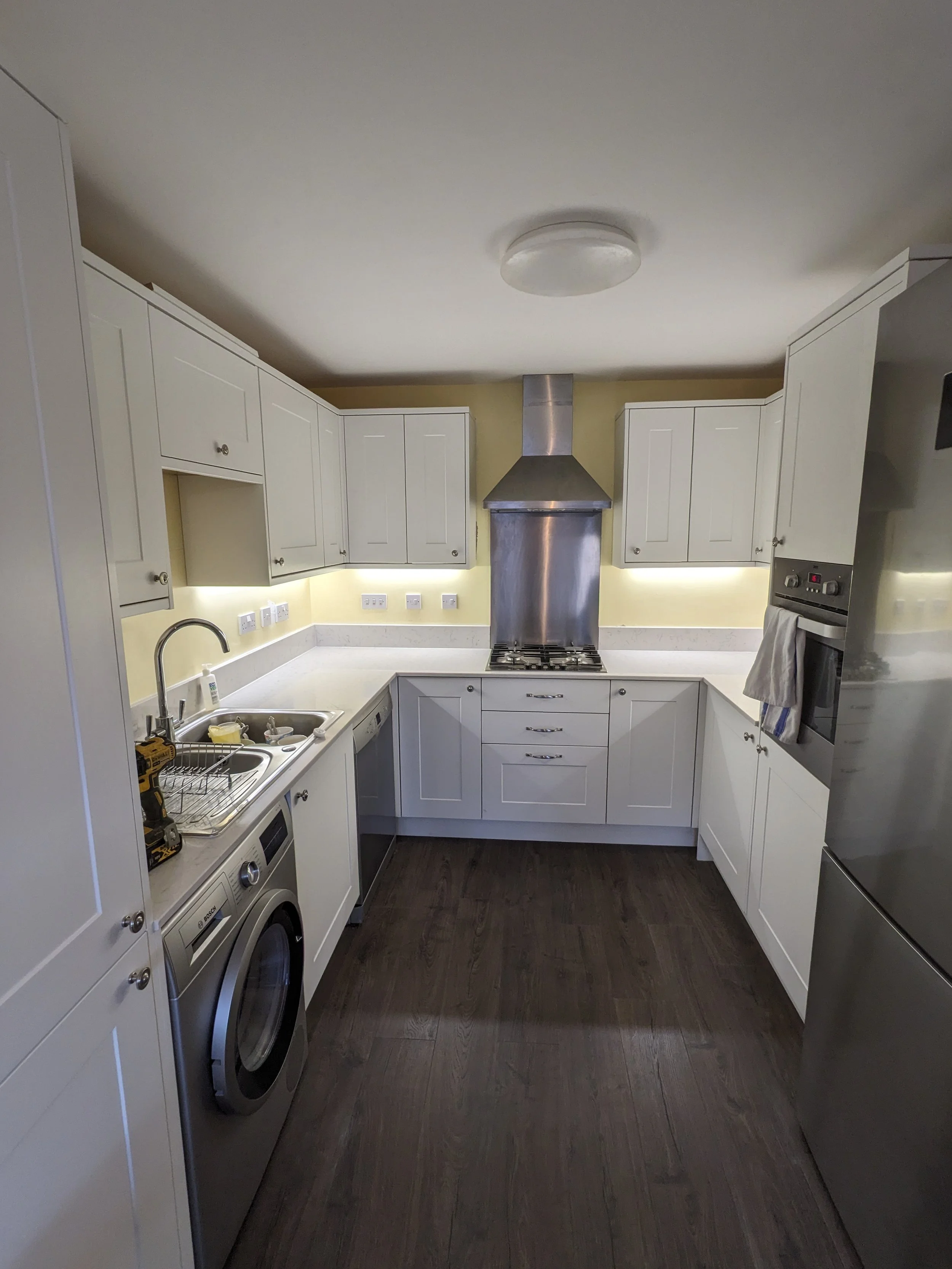 A newly installed kitchen with white cabinets, a stainless steel oven and fridge, and dark wooden flooring, with a washer under the sink and a stainless steel extractor hood above the stove.