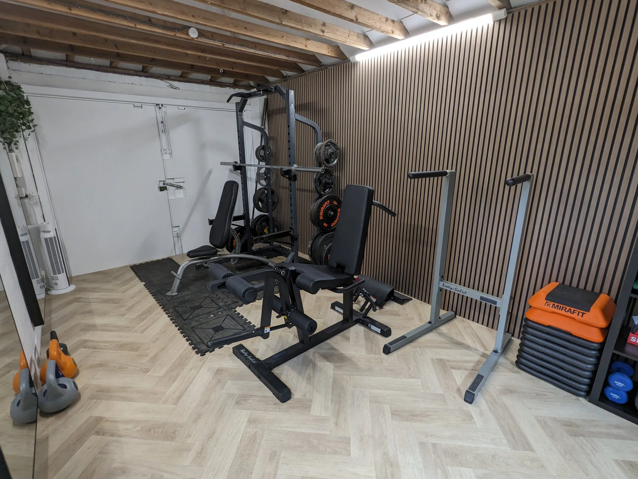 Home gym with weightlifting equipment including a weight bench, squat rack, and weight plates, along with kettlebells, resistance bands, and plyometric boxes.