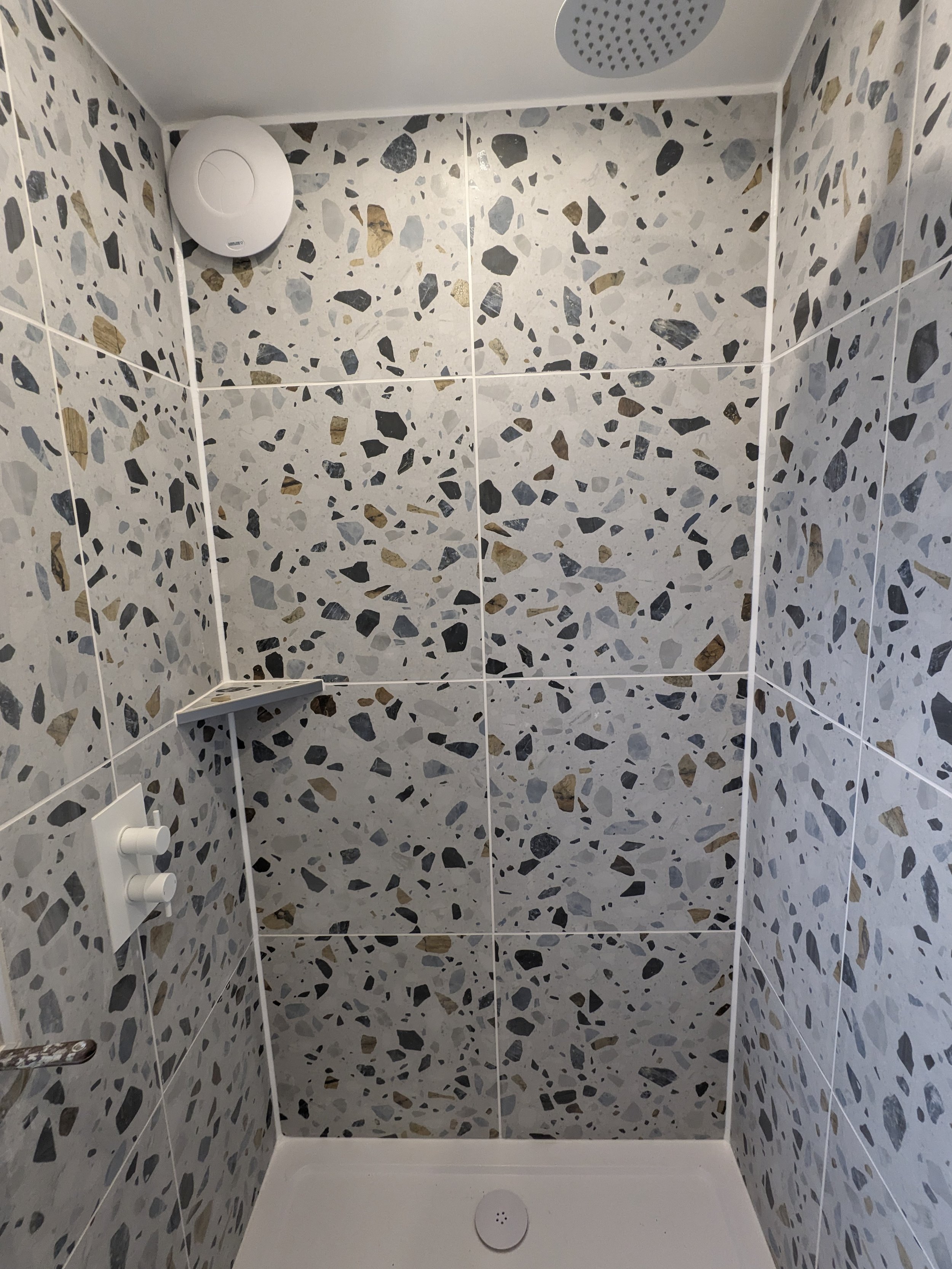 Interior of a shower with terrazzo tiles, a white shower drain, a soap shelf, an adjustable showerhead, and a control knob.