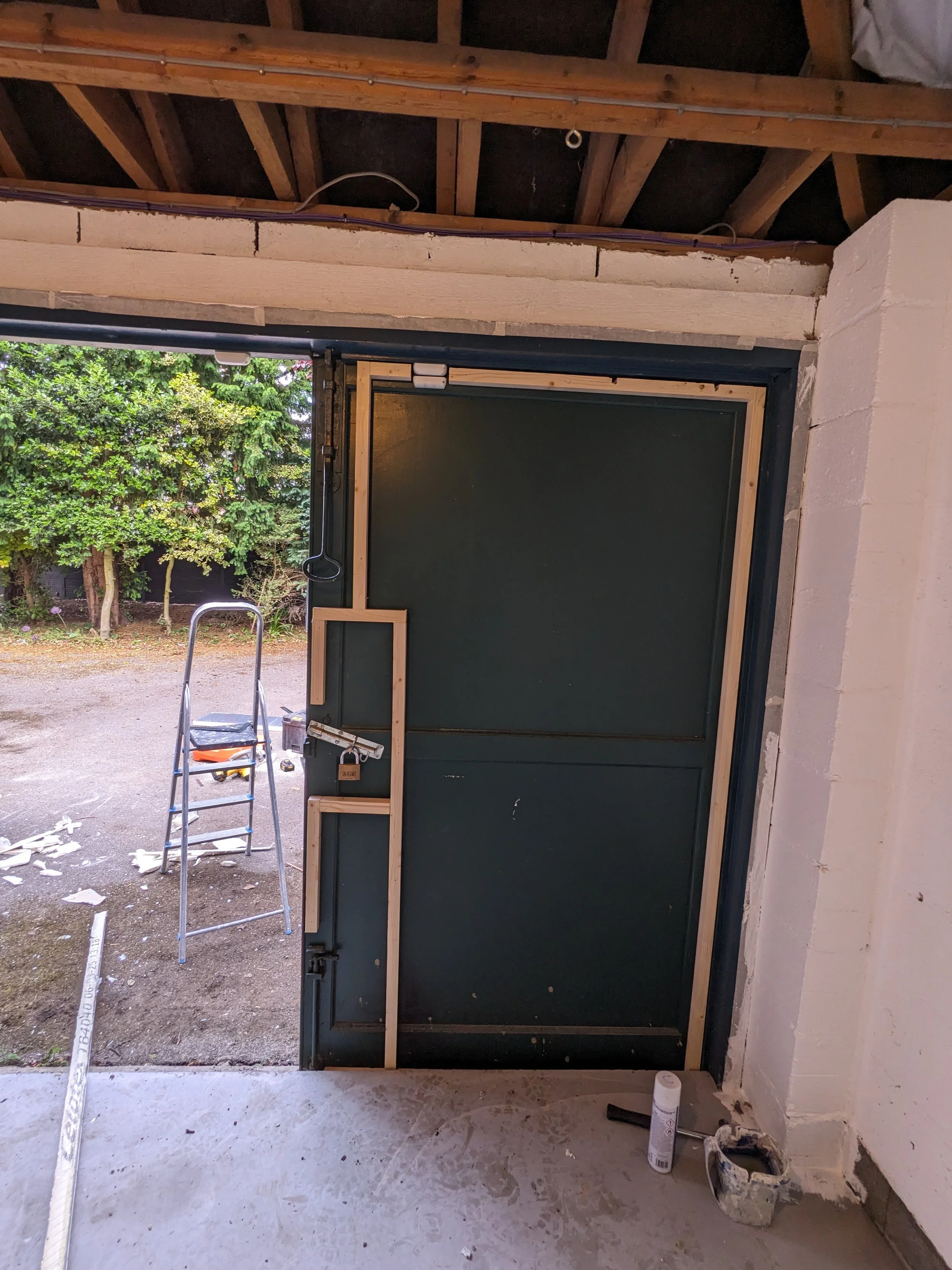 Interior view of a garage or workshop with a partially installed door frame, construction tools, and materials. Outer view shows a yard with trees and a dirt ground.