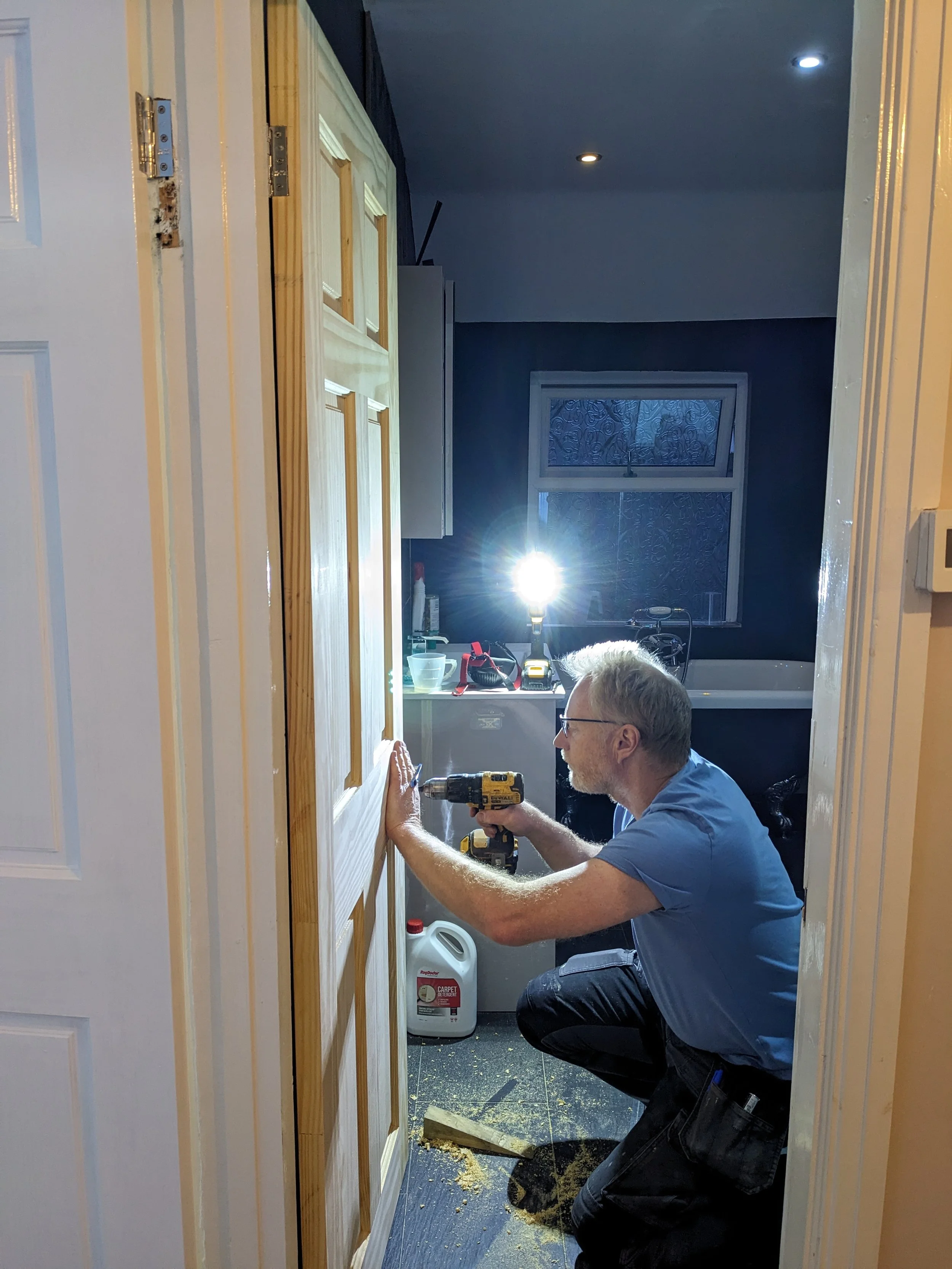 A man kneeling and working on a wooden door with a power drill in a laundry room, illuminated by a bright light.